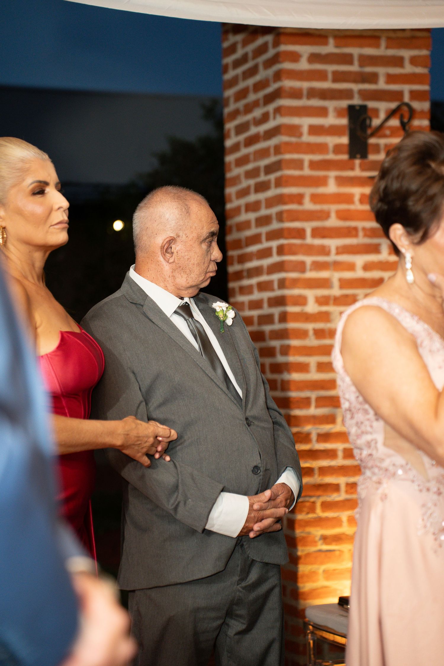 Elderly man in gray suit with boutonniere stands at wedding ceremony beside woman in red dress near brick wall.