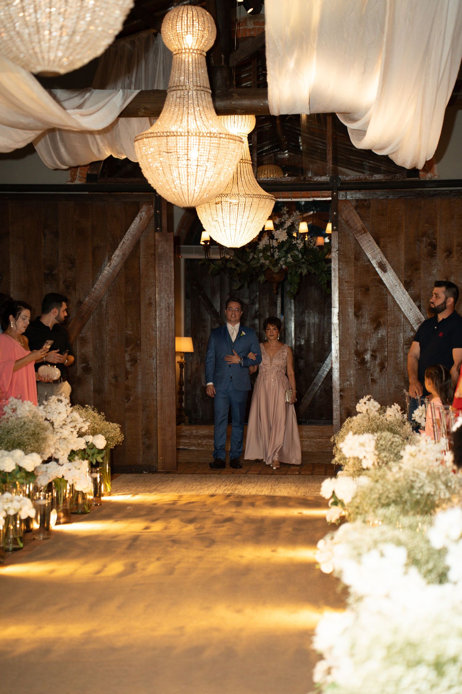 Elegant barn wedding ceremony with crystal chandeliers, floral aisle arrangements, and couple walking down the aisle.