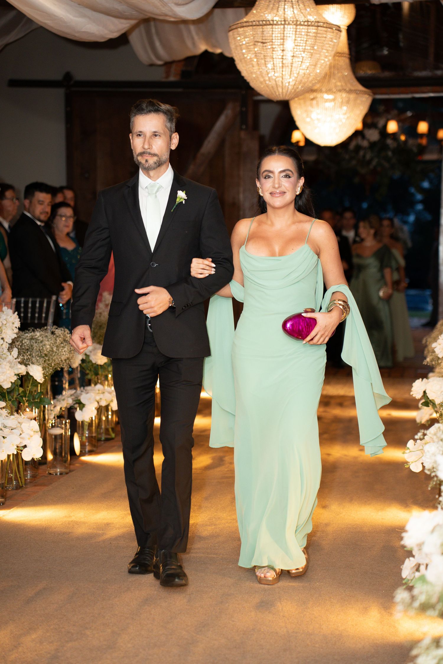 Elegant couple walking down wedding aisle, man in black suit, woman in mint green strapless gown holding purple clutch.
