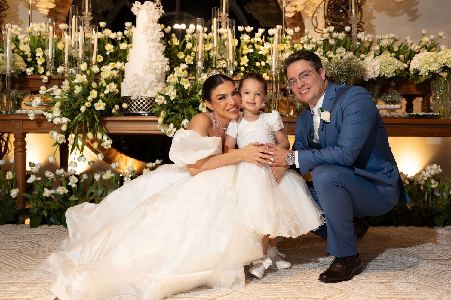 Bride in white gown and groom in blue suit pose with young flower girl in front of elegant floral wedding cake display.
