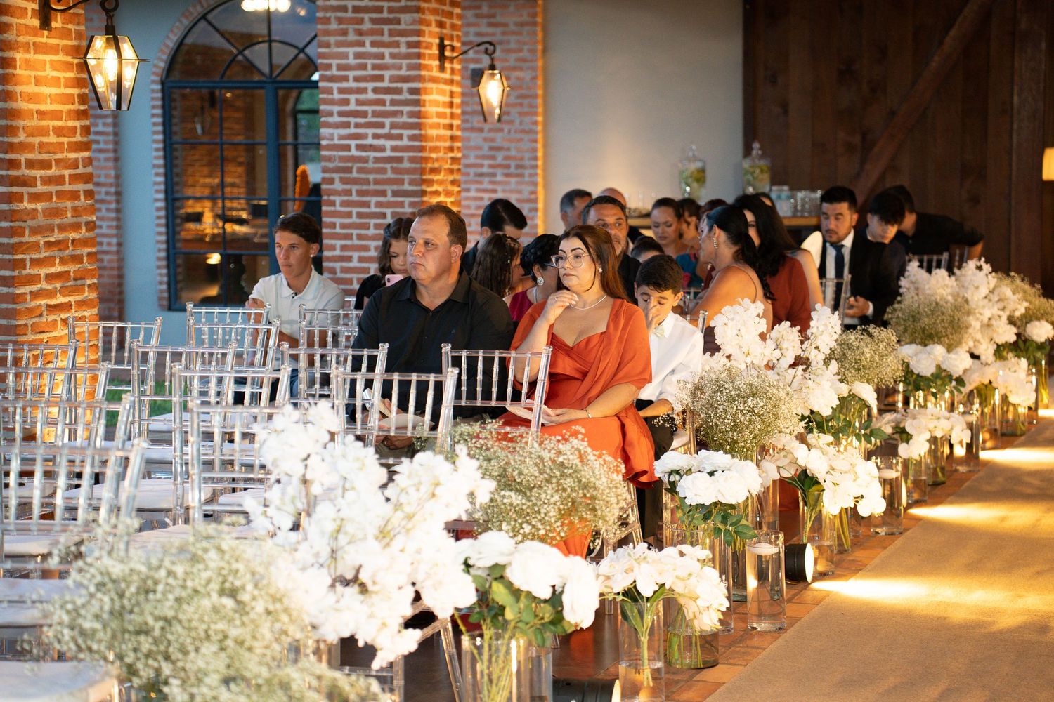 Elegant wedding ceremony with guests seated in white chairs beside lush white floral arrangements in a rustic brick venue.