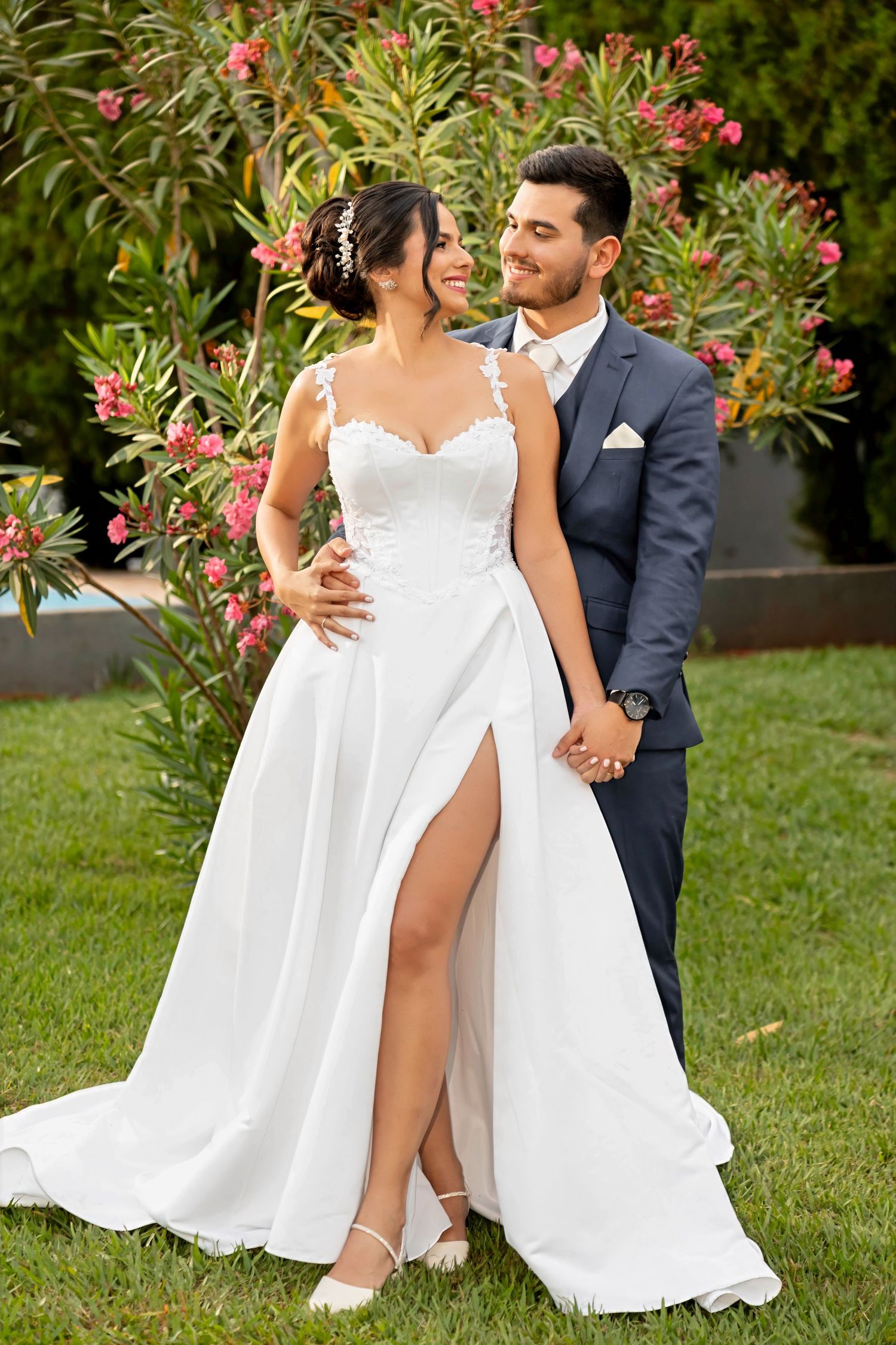 Bride in white lace gown with slit and groom in navy suit holding hands outdoors near flowering shrubs.