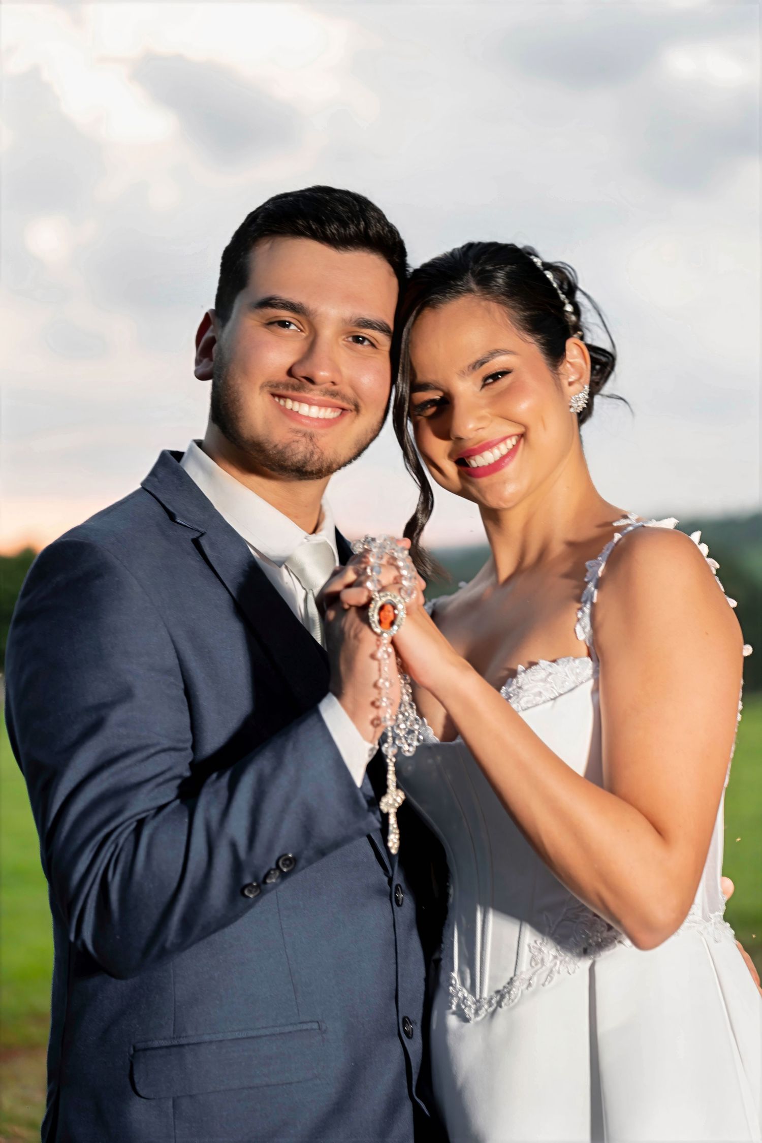 Smiling bride and groom holding a rosary together outdoors on their wedding day.