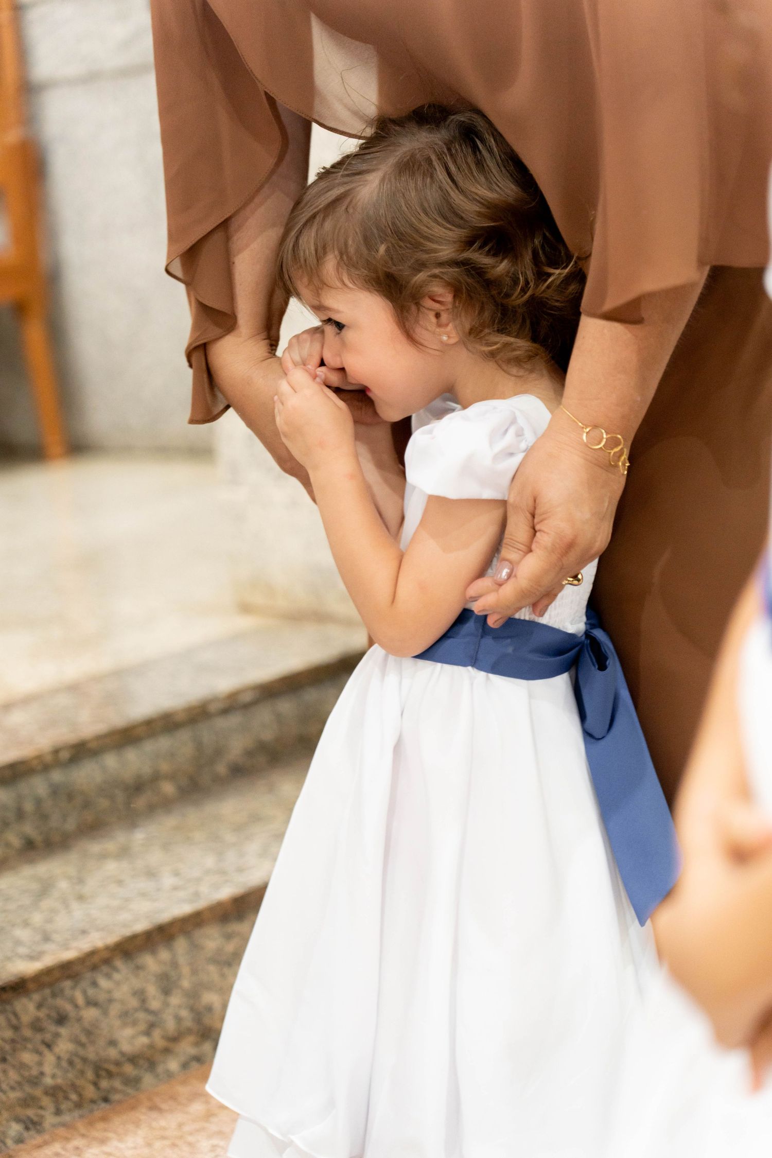 Emotional flower girl in white dress with blue bow cries while clinging to an adult during a wedding ceremony.