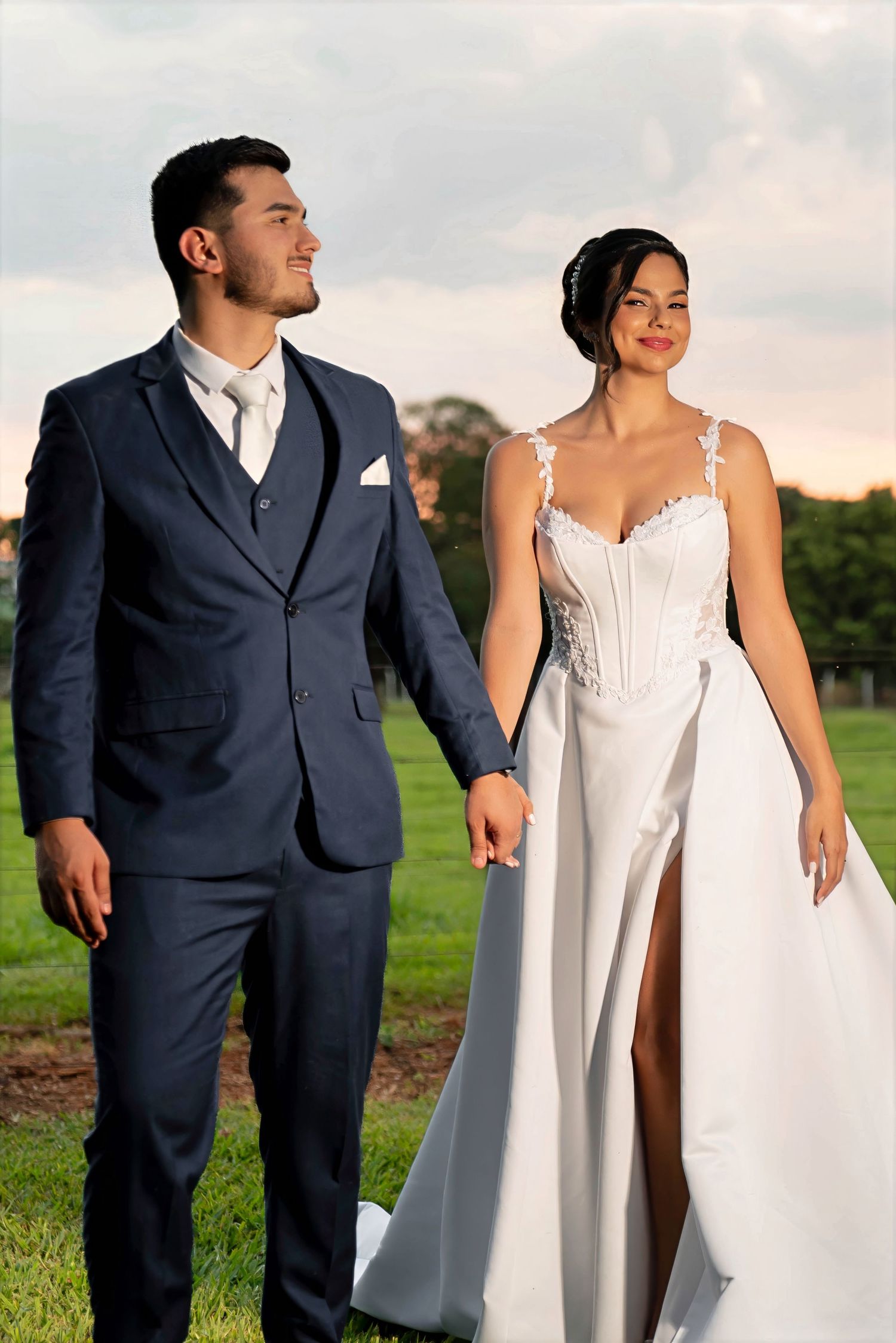 A couple in wedding attire holds hands outdoors, groom in navy suit, bride in white gown with lace details.