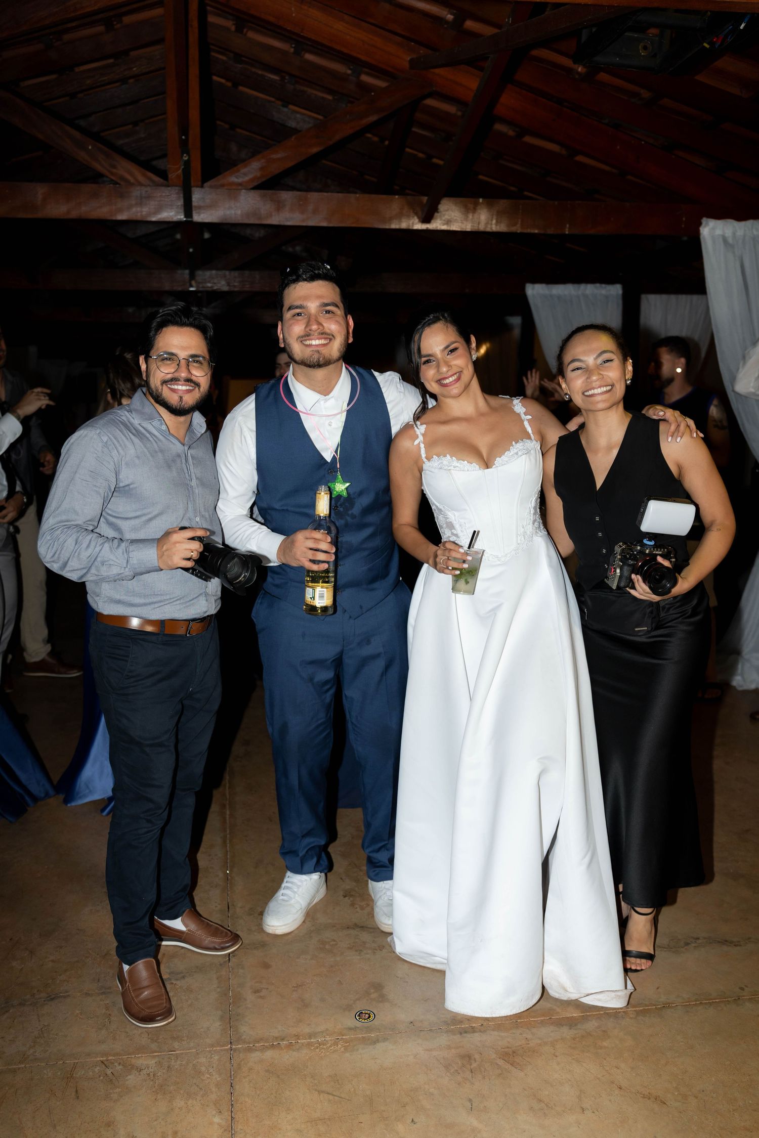 Four guests pose together at a wedding reception, including a bride in a white gown, inside a rustic wood-beamed venue.