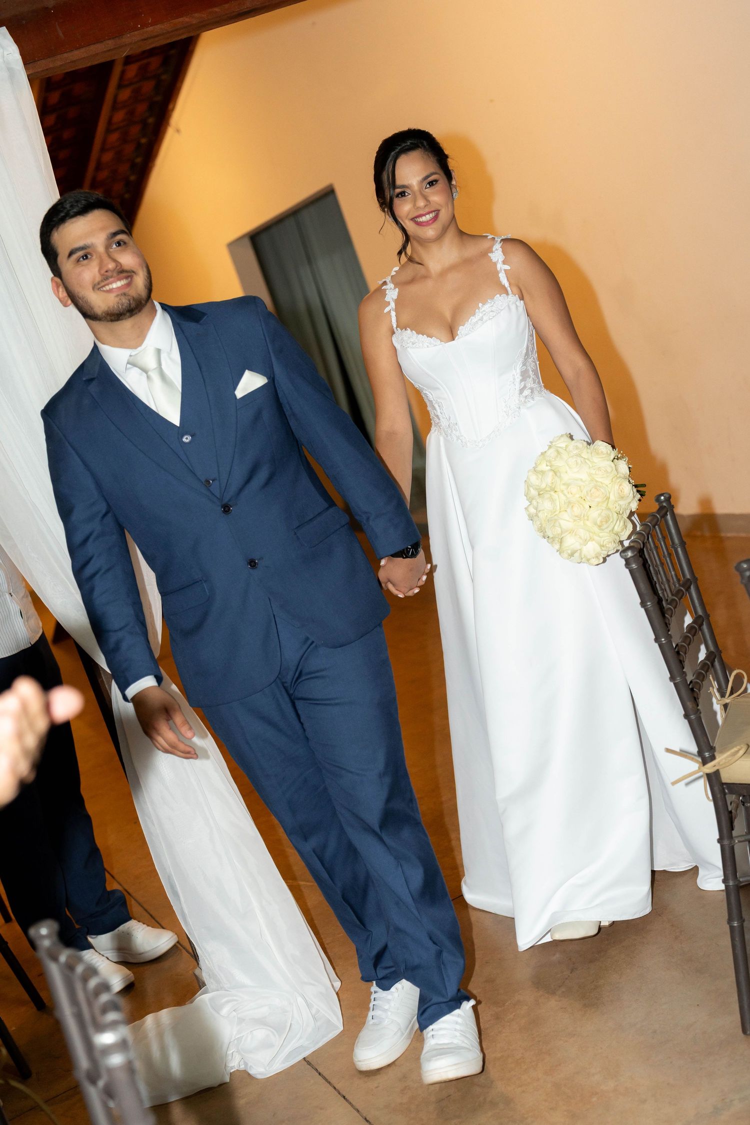 A smiling couple walks hand in hand at their wedding, groom in navy suit, bride in white gown holding a bouquet.