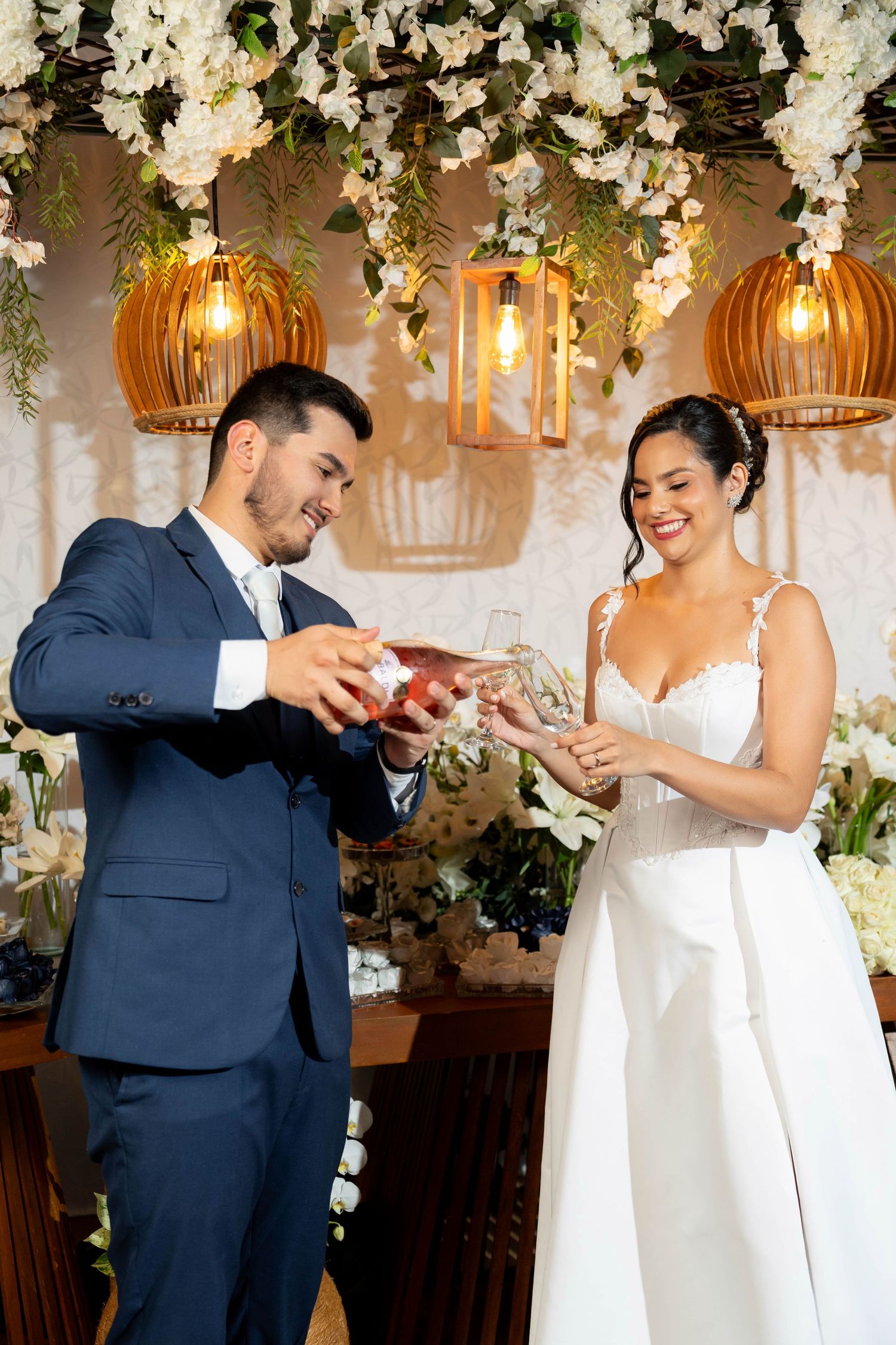 Smiling bride and groom exchanging rings at elegant wedding ceremony with floral decorations and warm pendant lights.