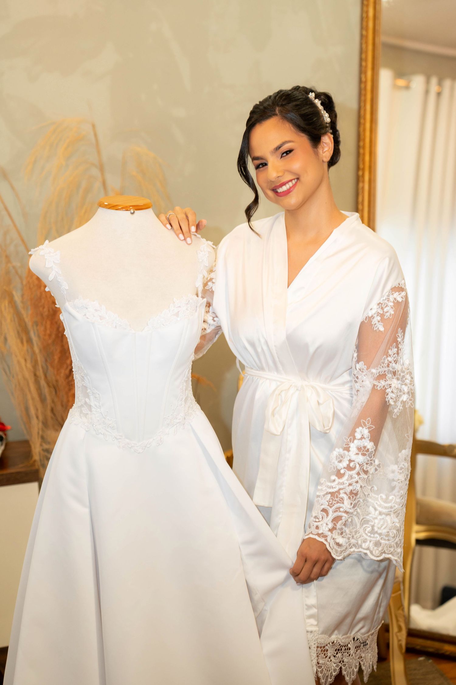 Smiling bride in white lace robe stands beside a mannequin displaying an elegant off-shoulder wedding gown in a bridal boutique.