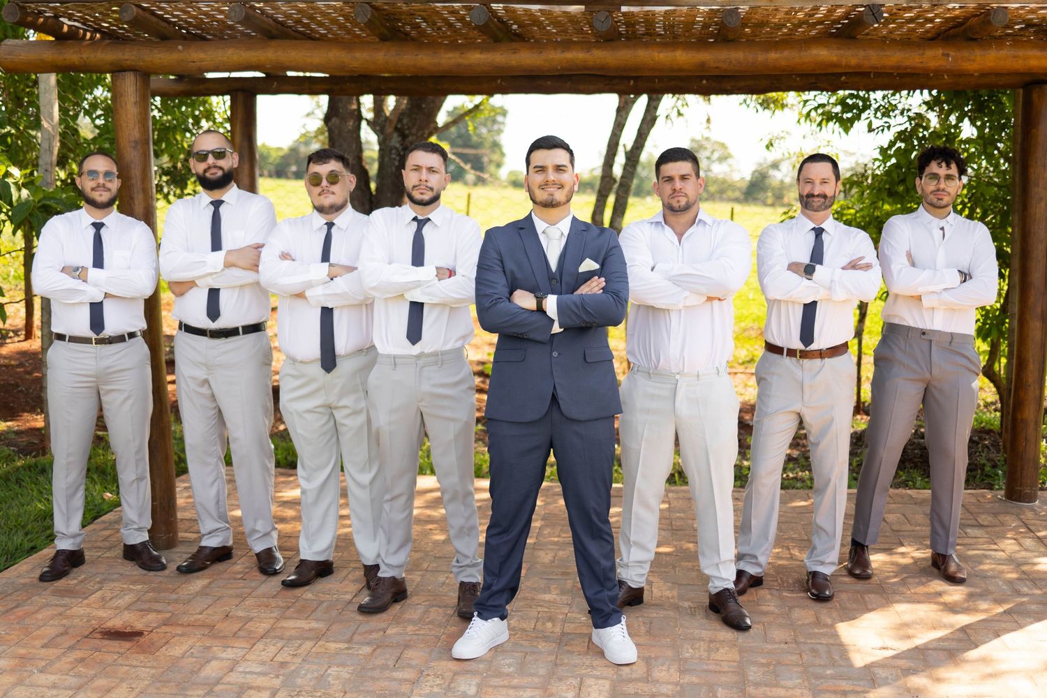 Groom in navy suit stands confidently with groomsmen in matching gray pants and white shirts with black ties outdoors.