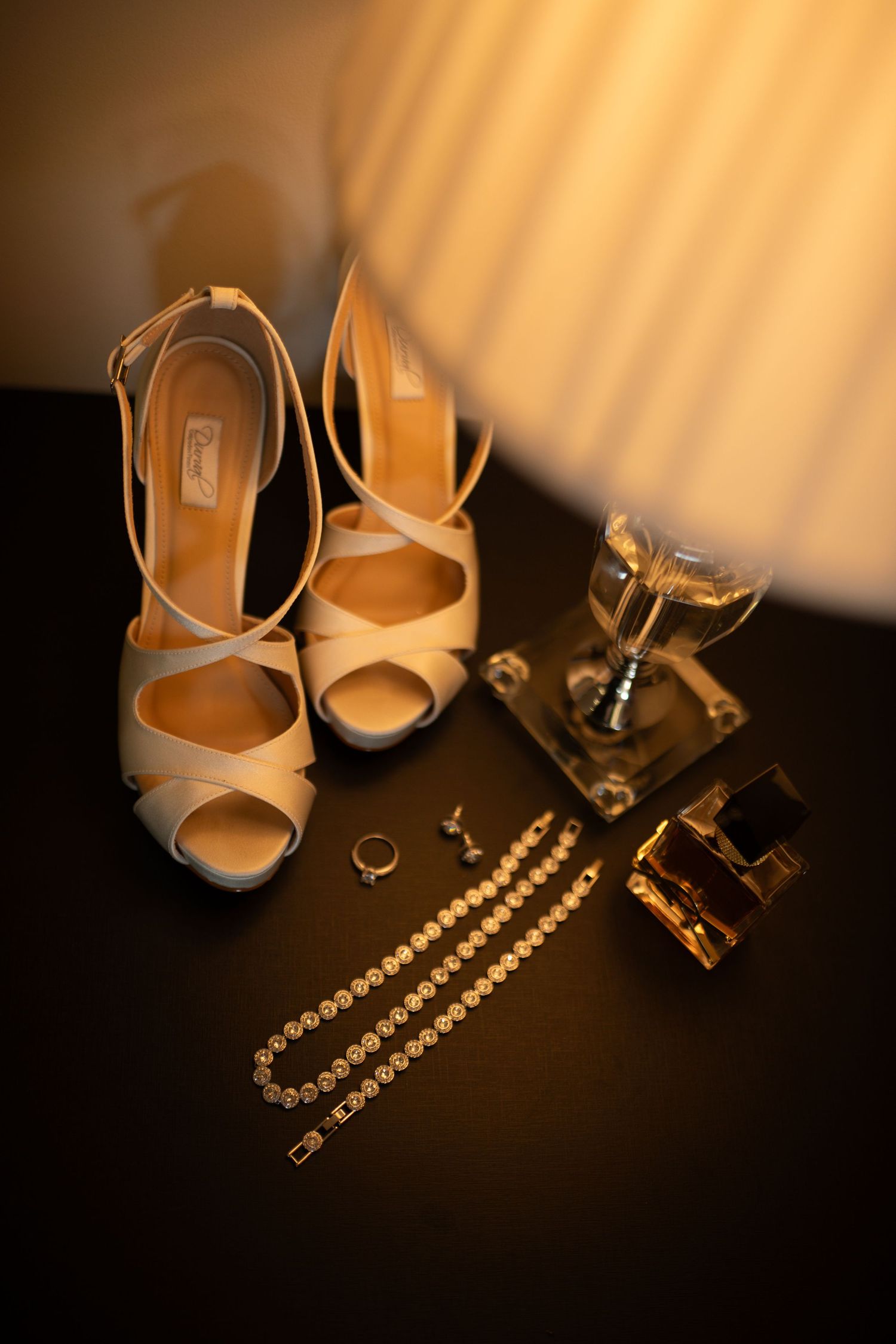 Elegant bridal accessories including nude heels, diamond bracelet, gold ring, earrings, and perfume bottles on dark surface.