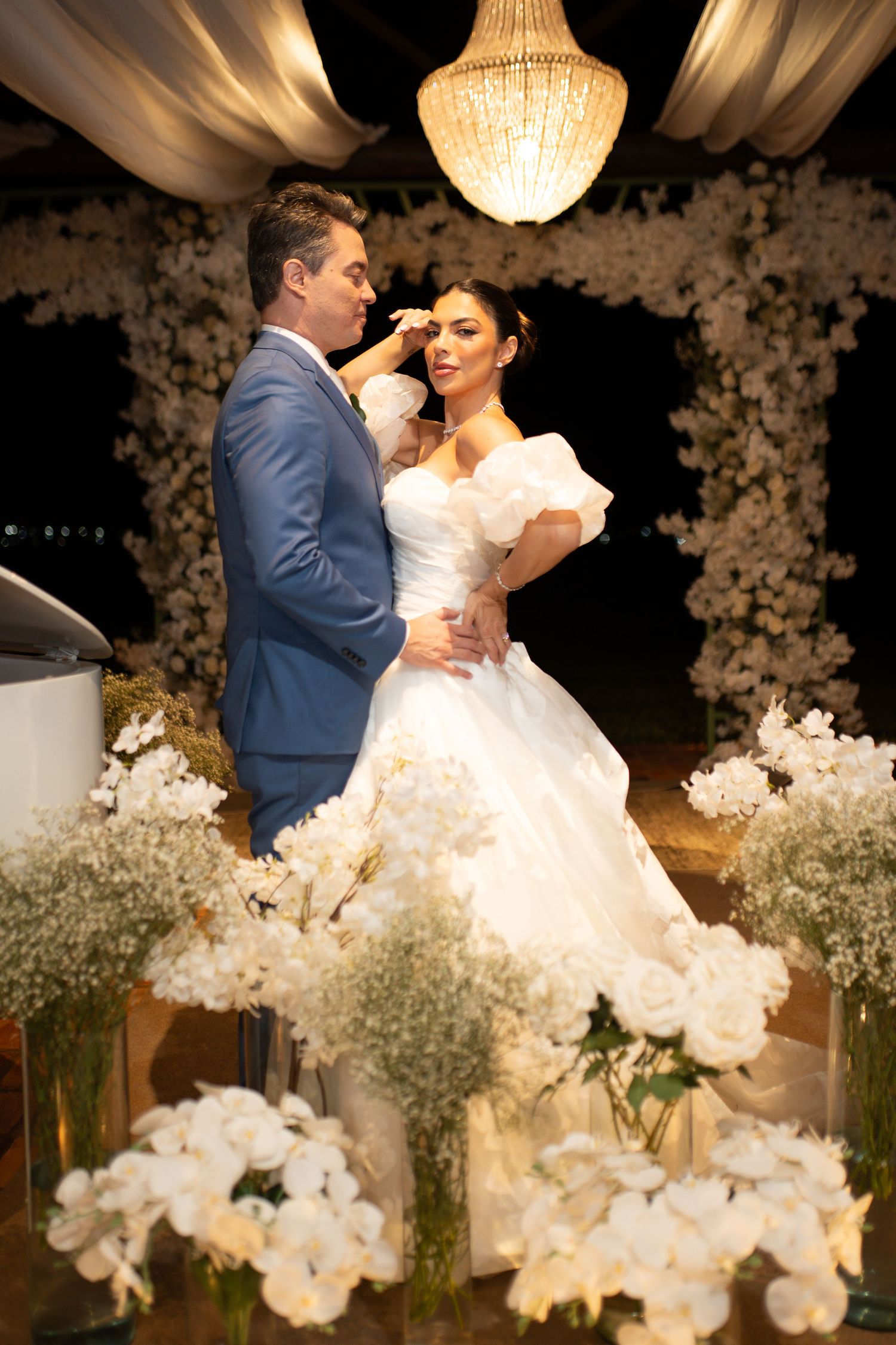Bride and groom share a romantic moment under a chandelier surrounded by white floral arrangements at an elegant wedding ceremony.