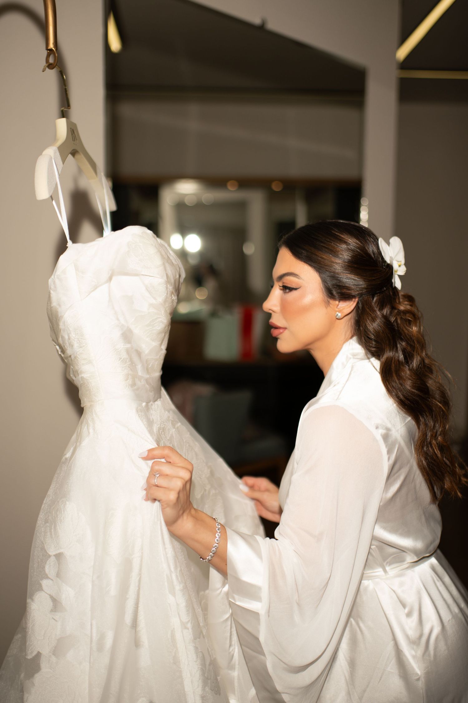 Bride in white robe admiring her wedding dress on a mannequin in a bridal boutique.