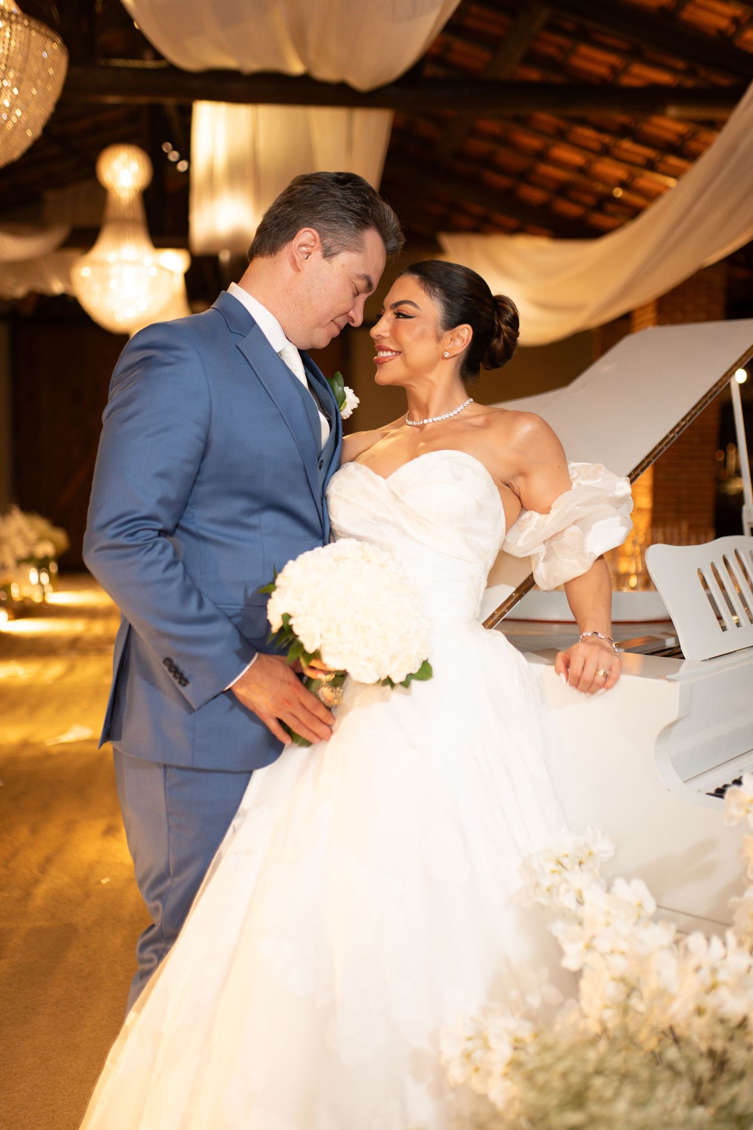 Bride in white ball gown and groom in blue suit share romantic moment at elegant indoor wedding venue.