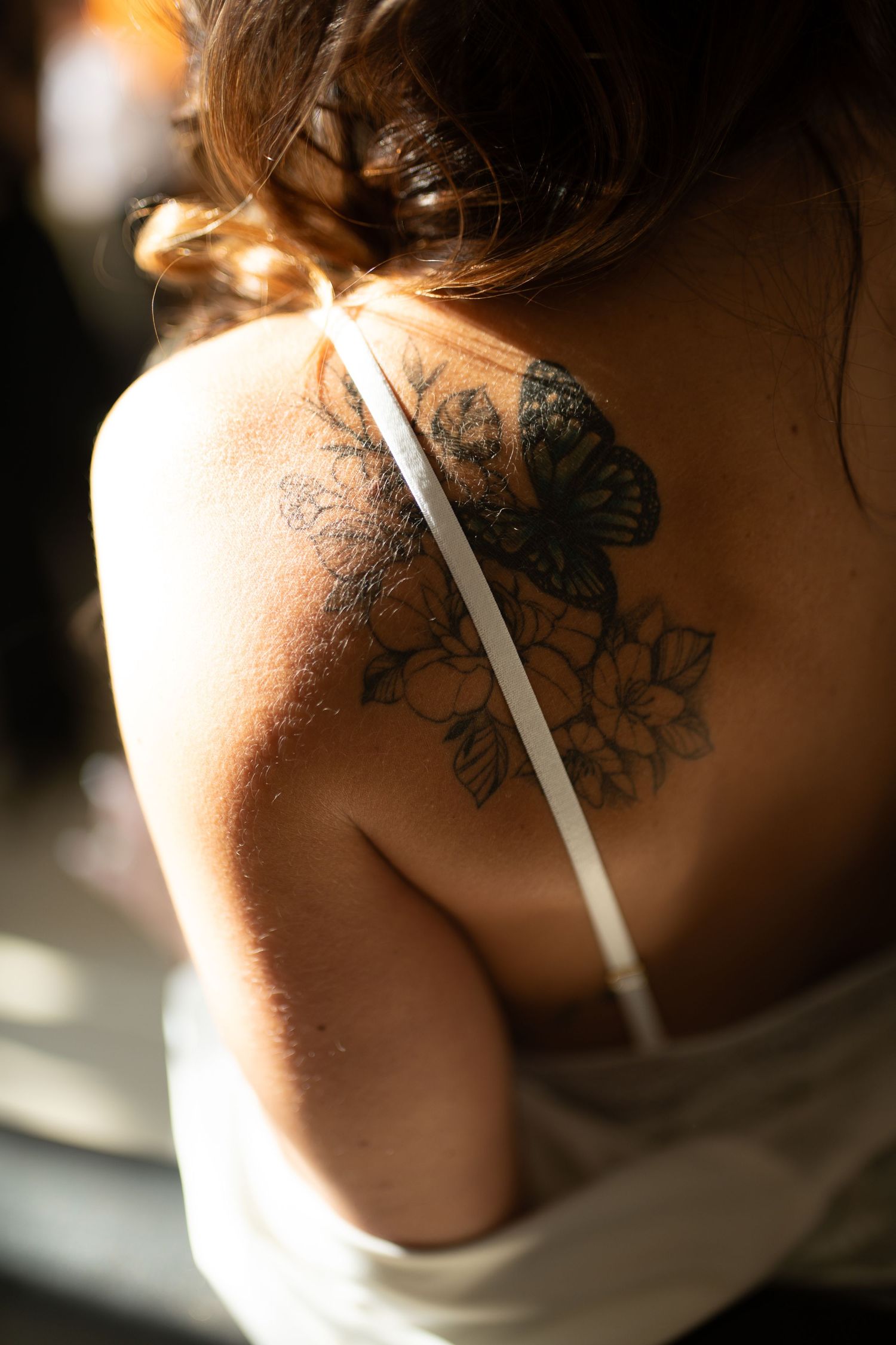 Woman's back tattoo featuring a butterfly and floral design, visible over a white dress strap in natural sunlight.
