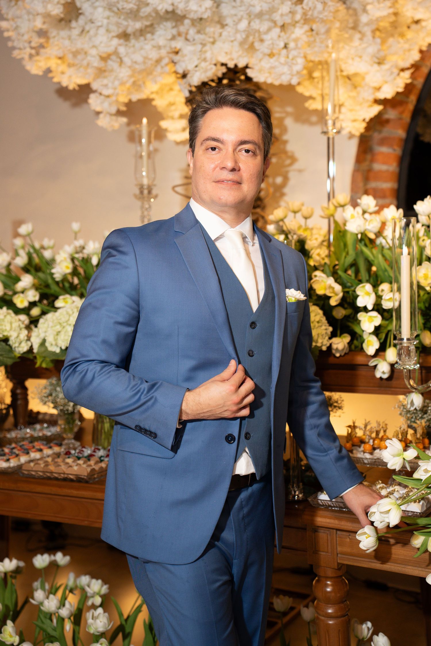 Man in blue three-piece suit poses elegantly at a floral wedding reception with white flowers and candlelight.