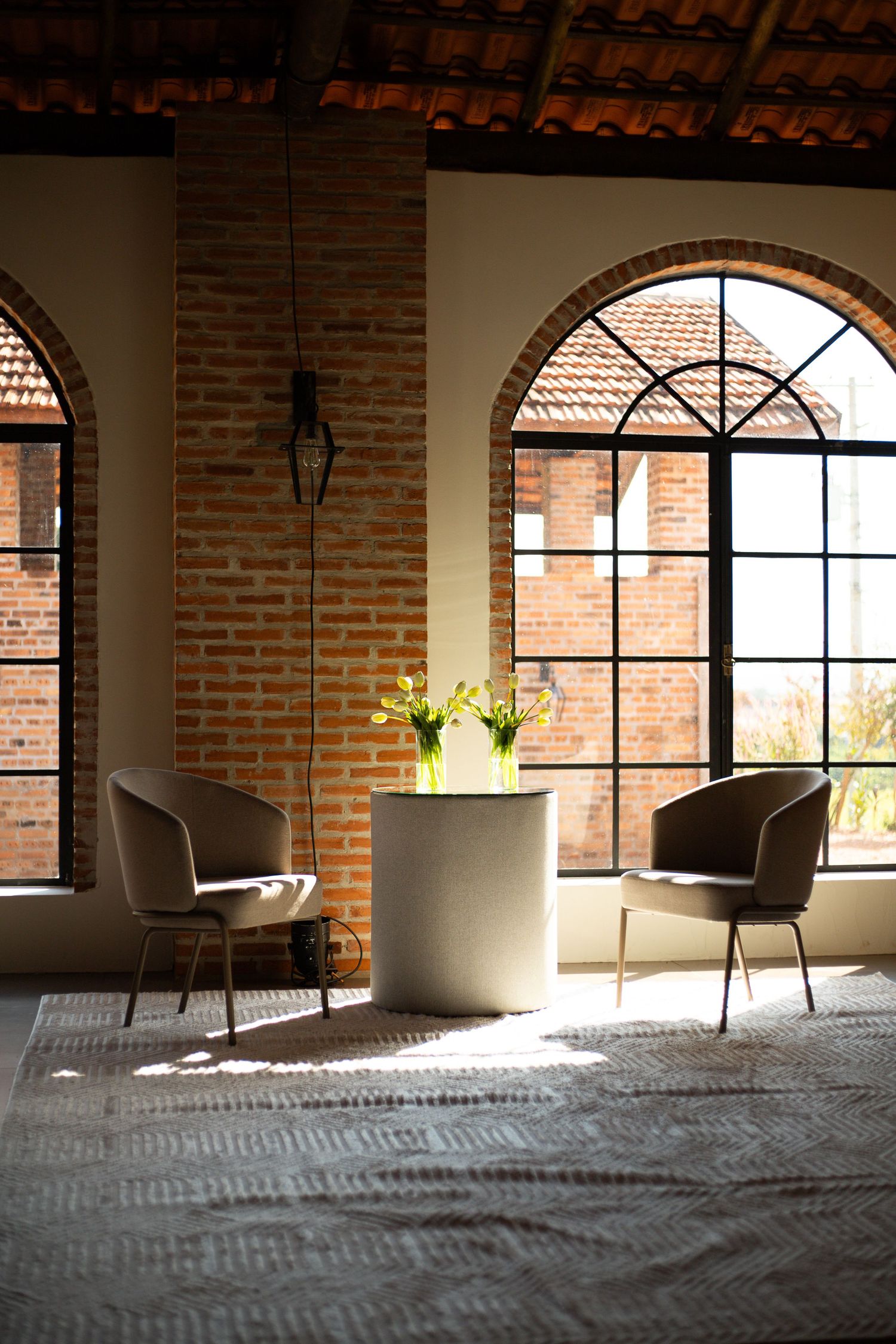 Elegant industrial lounge with arched windows, brick walls, two gray chairs, and a round table with yellow flowers.