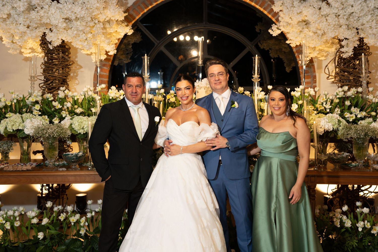 Elegant wedding reception with bride in white gown and groom in blue suit posing with two guests amid floral decor.