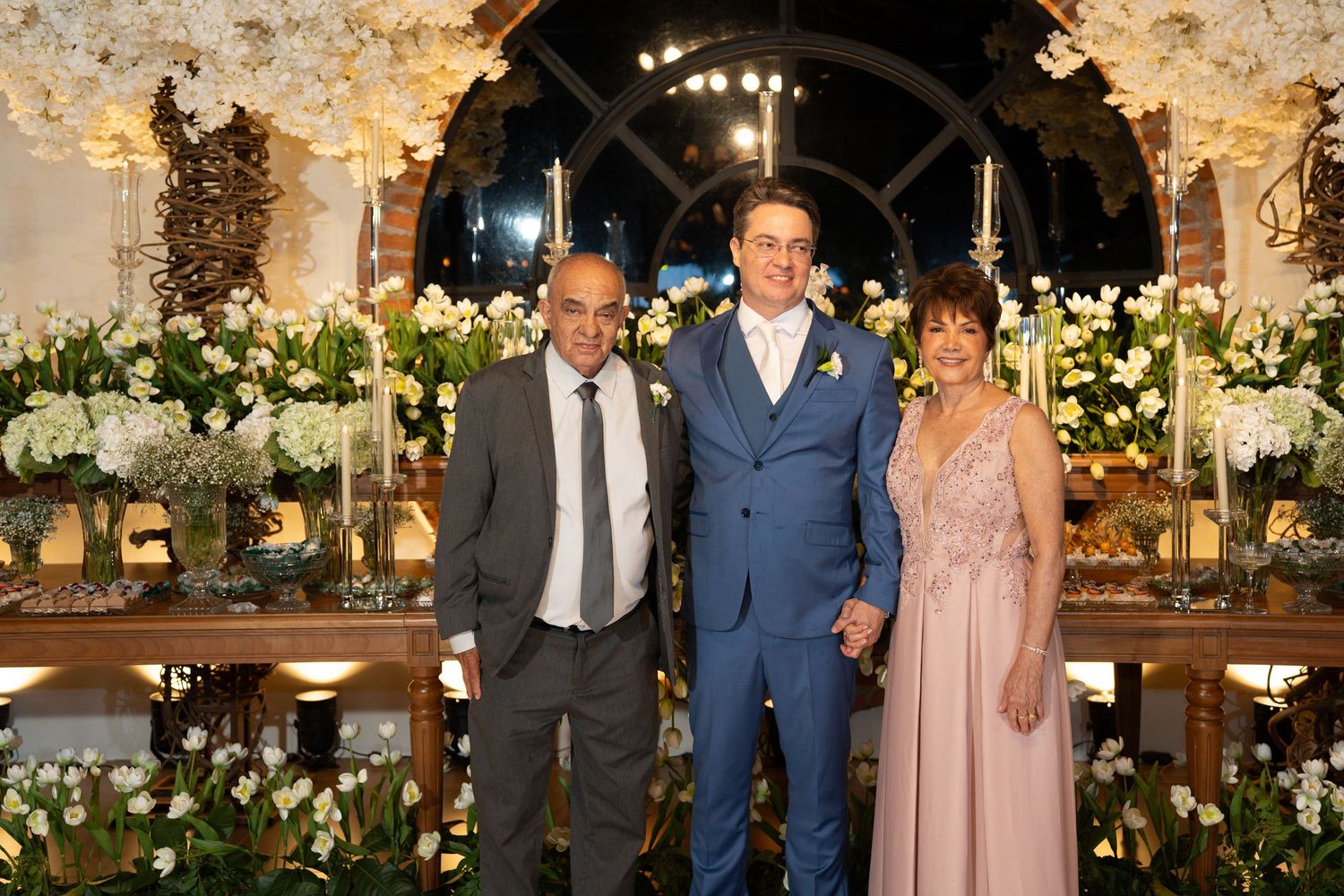 A groom in a blue suit poses with two guests at an elegant floral wedding reception.