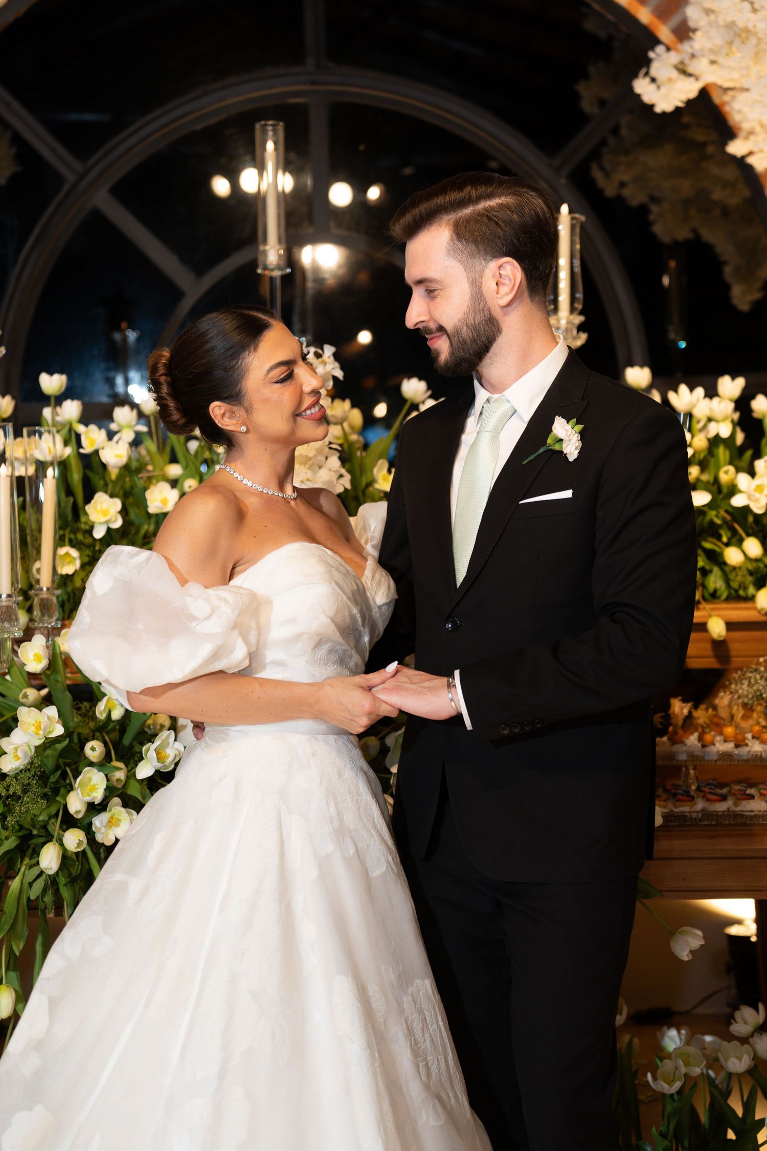Bride and groom holding hands at elegant wedding ceremony with white tulips and floral arch backdrop.