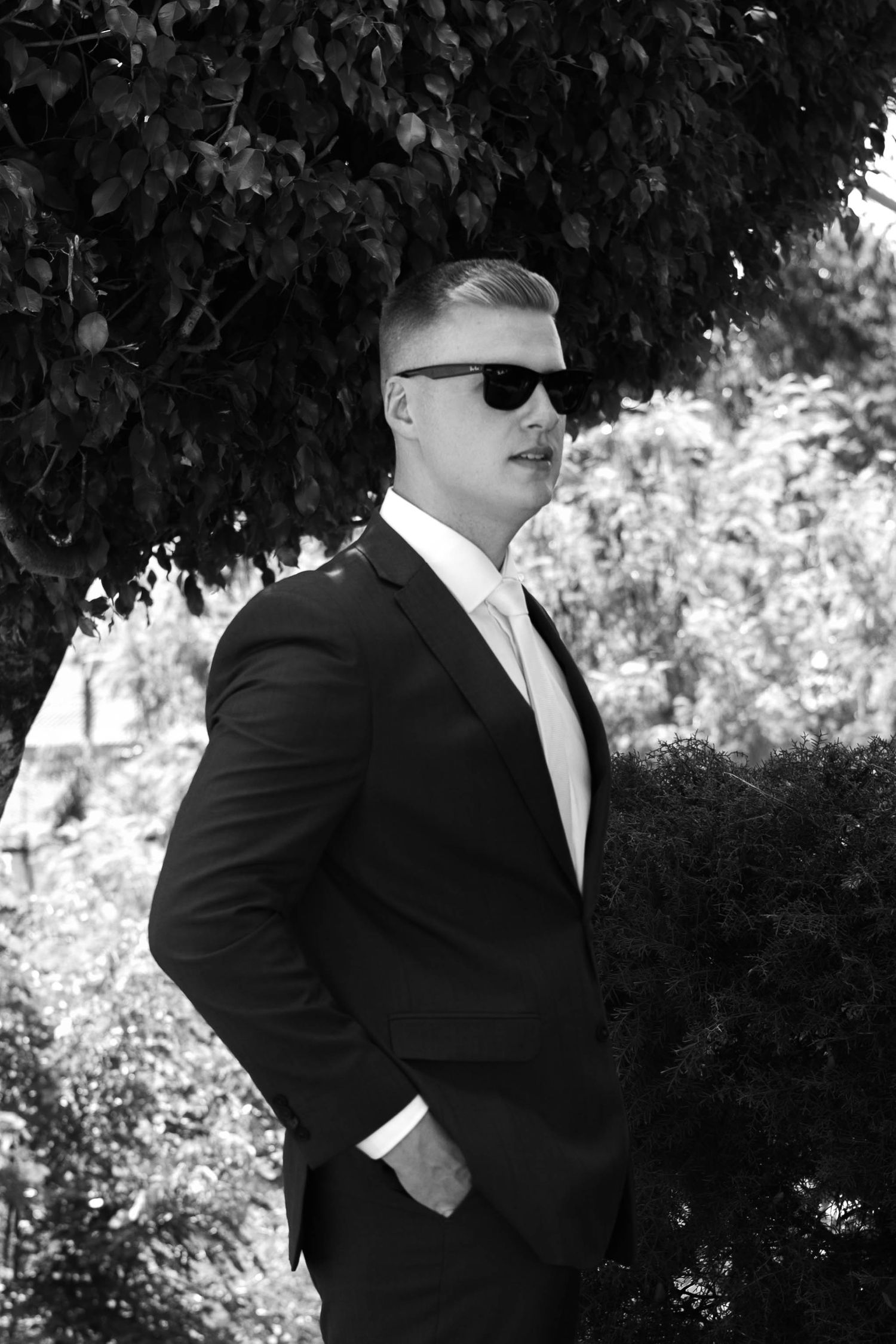 Man in black suit and sunglasses posing outdoors among lush green trees in a black and white photo.