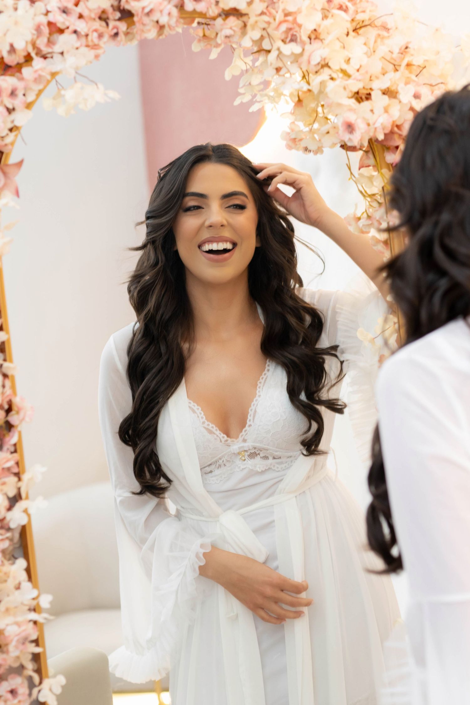 Laughing bride in white lace robe getting hair styled near floral arch mirror.