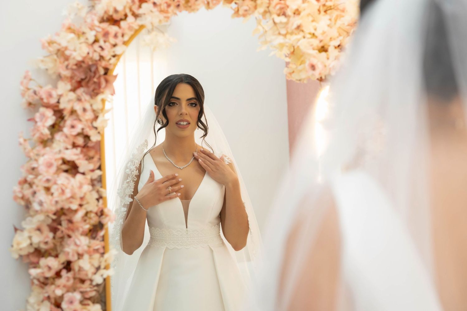 Bride in white ball gown wedding dress with veil stands before floral arch mirror in bridal boutique fitting room.