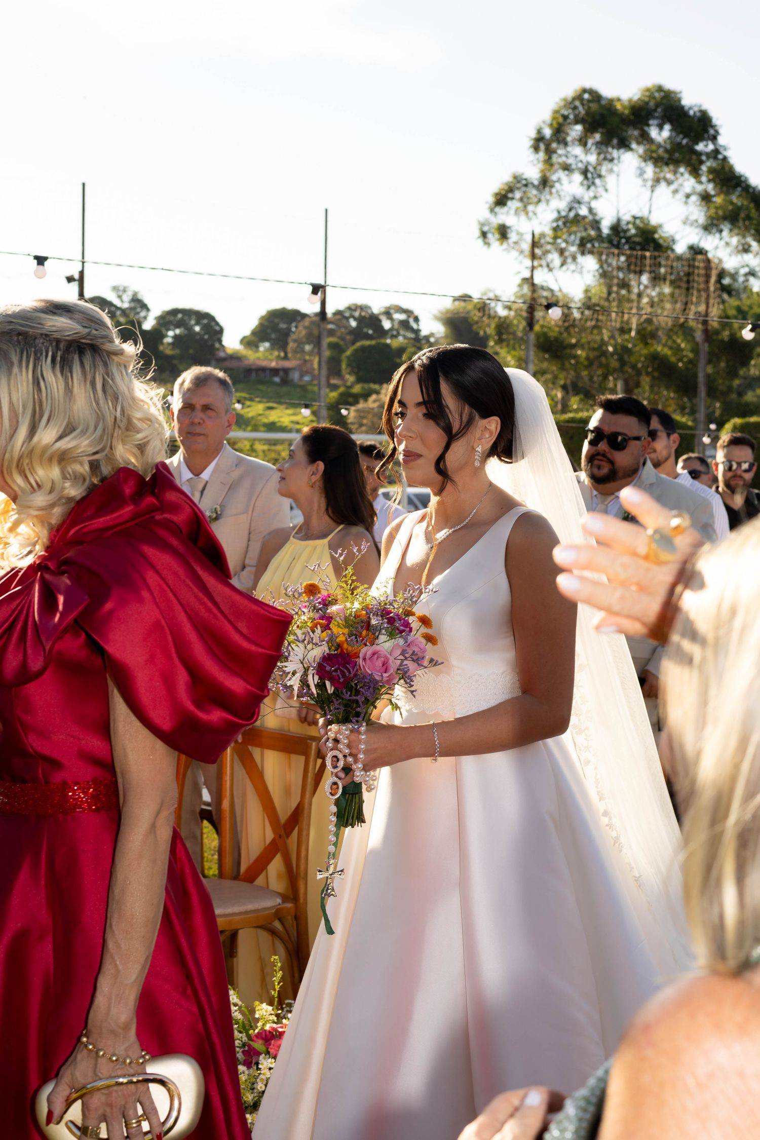 Bride in white gown holding colorful bouquet greeted by guest in red dress at outdoor wedding ceremony.