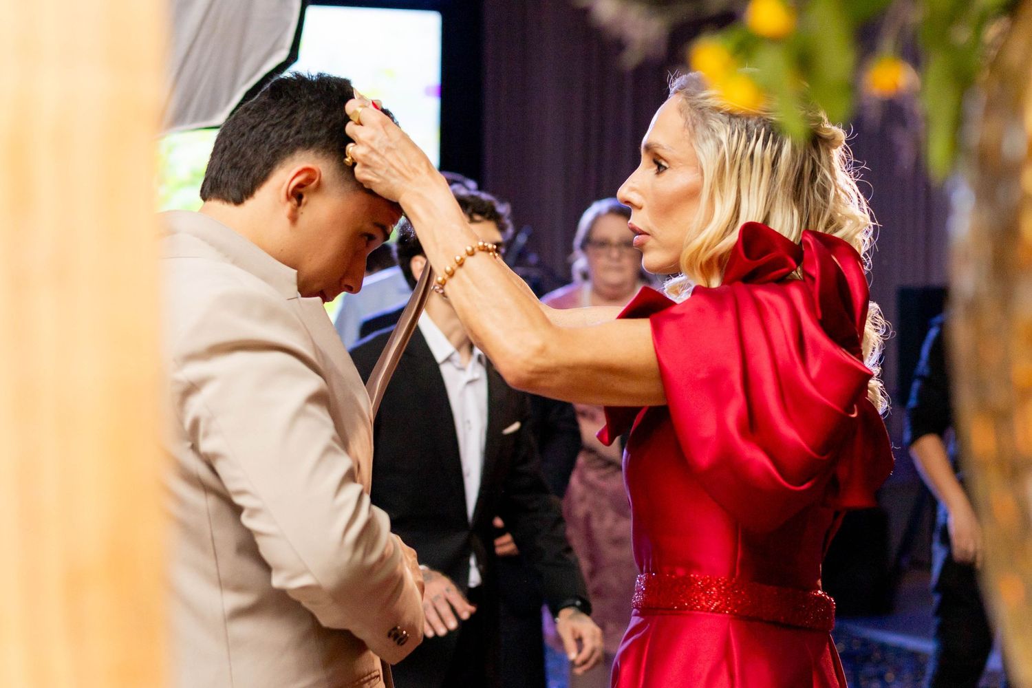 Woman in red dress placing bracelet on bowing man in beige suit at elegant indoor event.