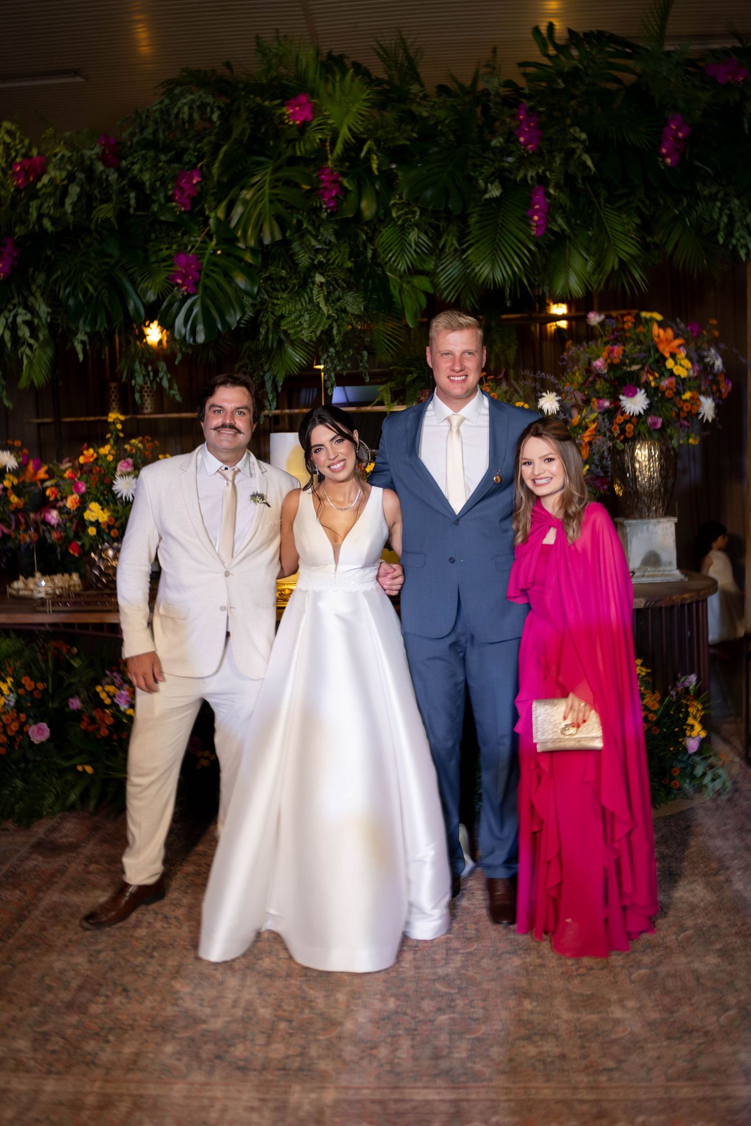 Wedding party of four posing with bride in white gown, groom in blue suit, and guests in cream and pink attire.