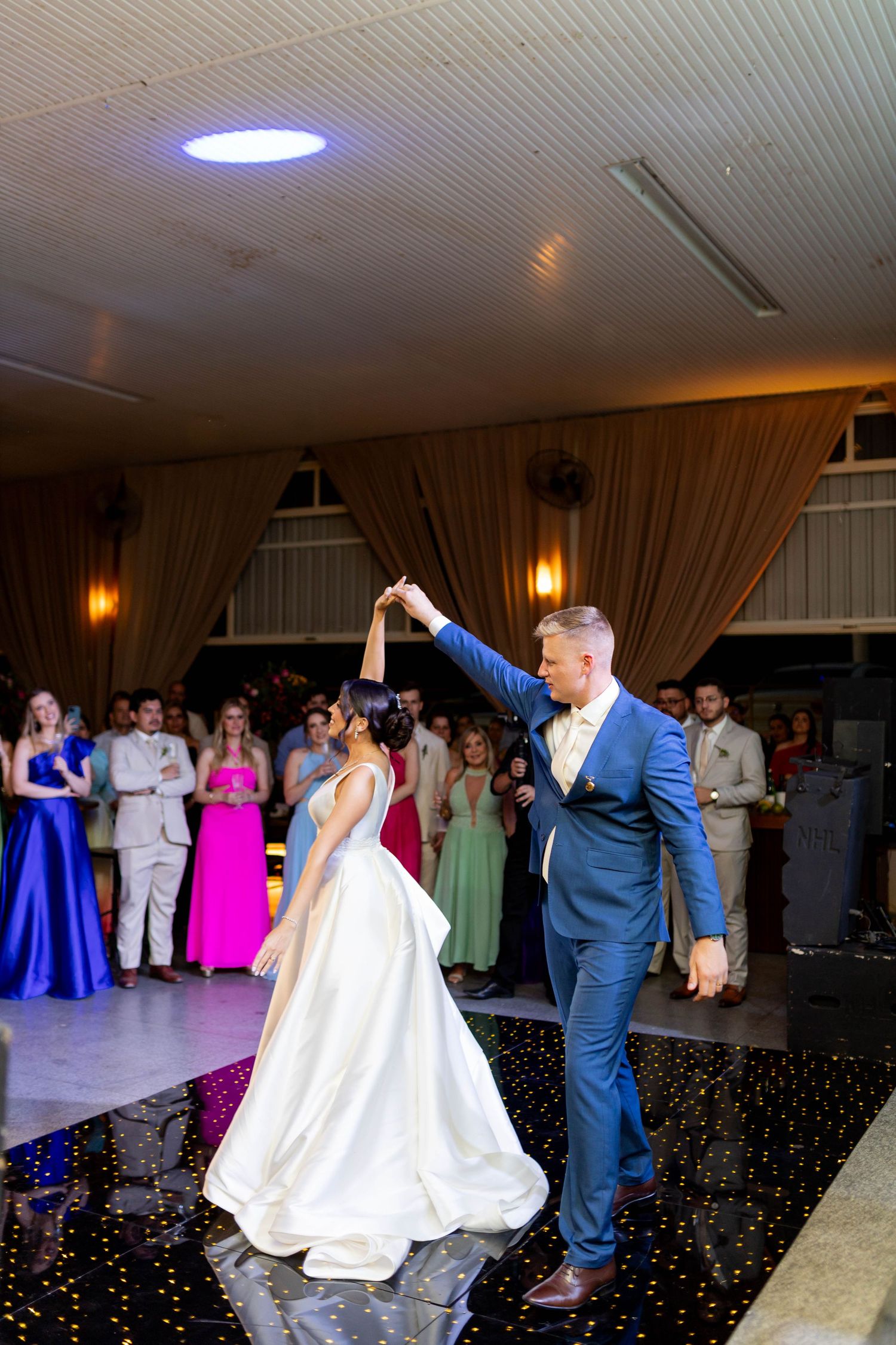 Bride and groom share their first dance at wedding reception while guests watch in colorful formal attire.