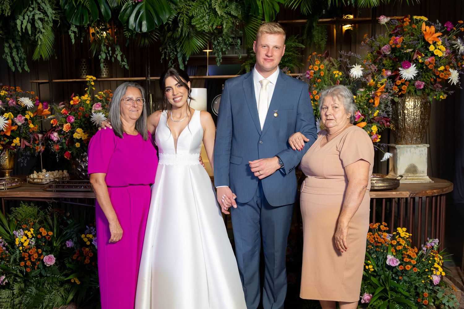 Wedding couple in formal attire poses with two older women amid colorful floral backdrop at elegant reception venue.