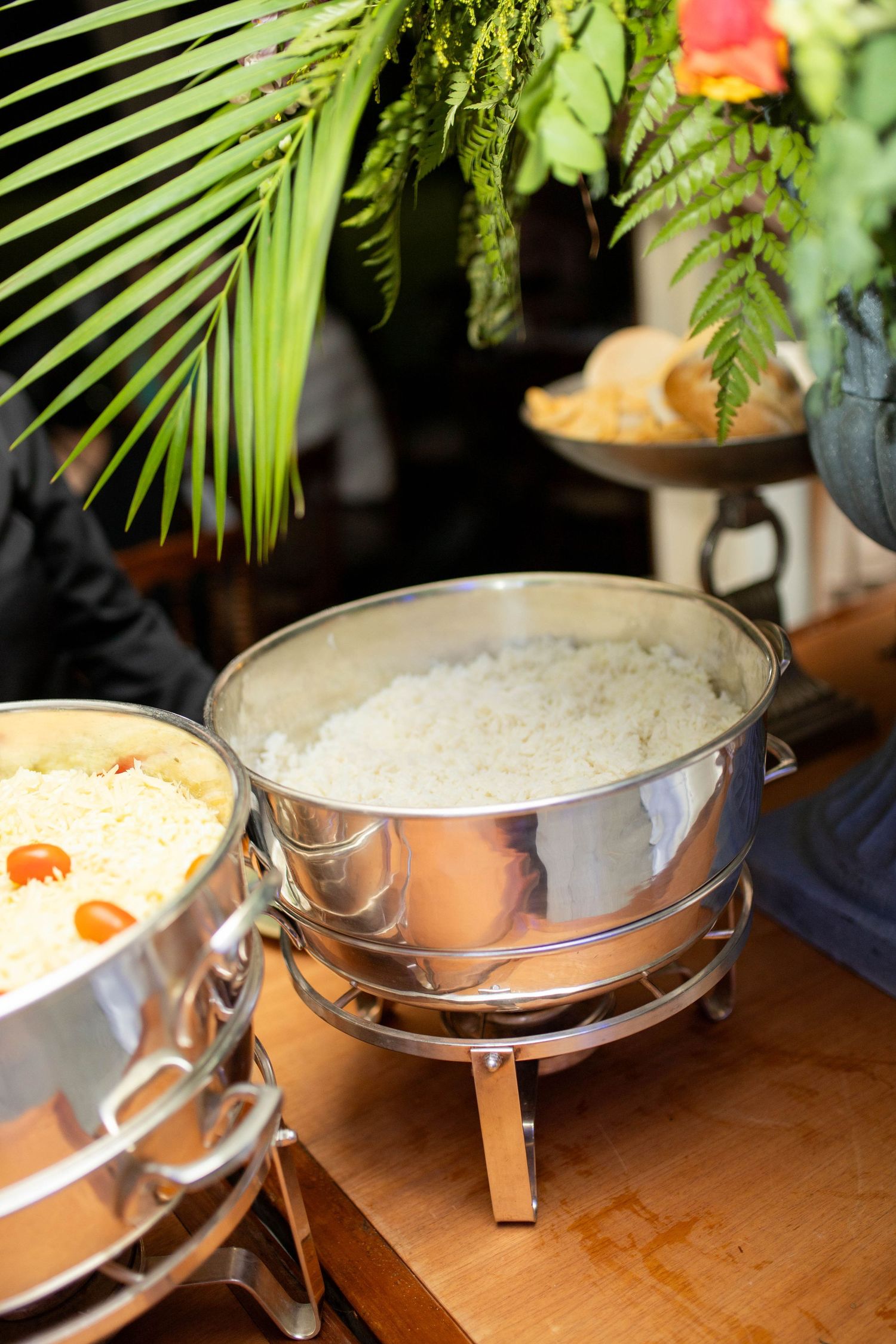 Stainless steel chafing dishes with rice and yellow curry on a wooden buffet table, surrounded by tropical green plants.