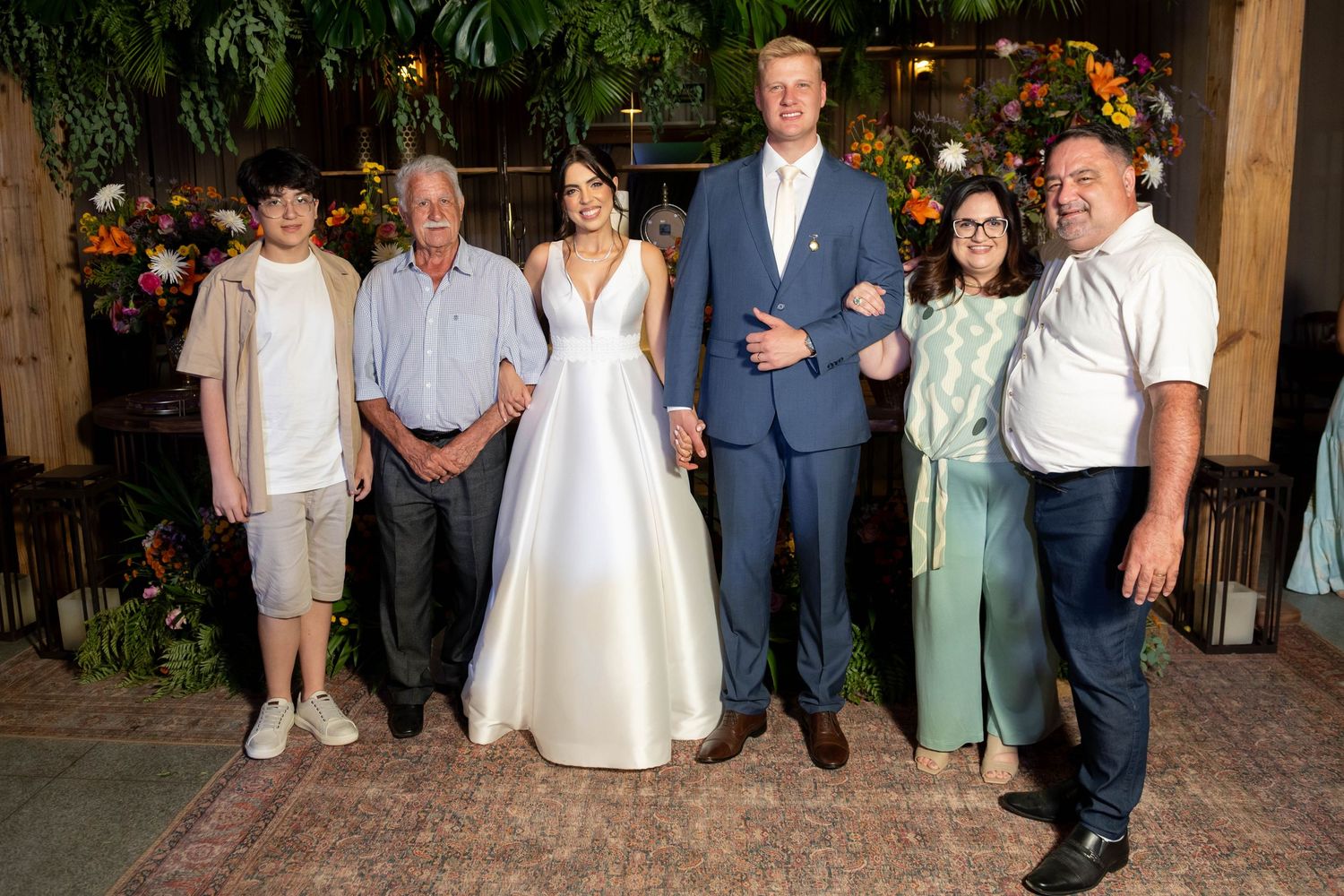 Wedding couple in formal attire poses with four family members indoors amid tropical floral decor.