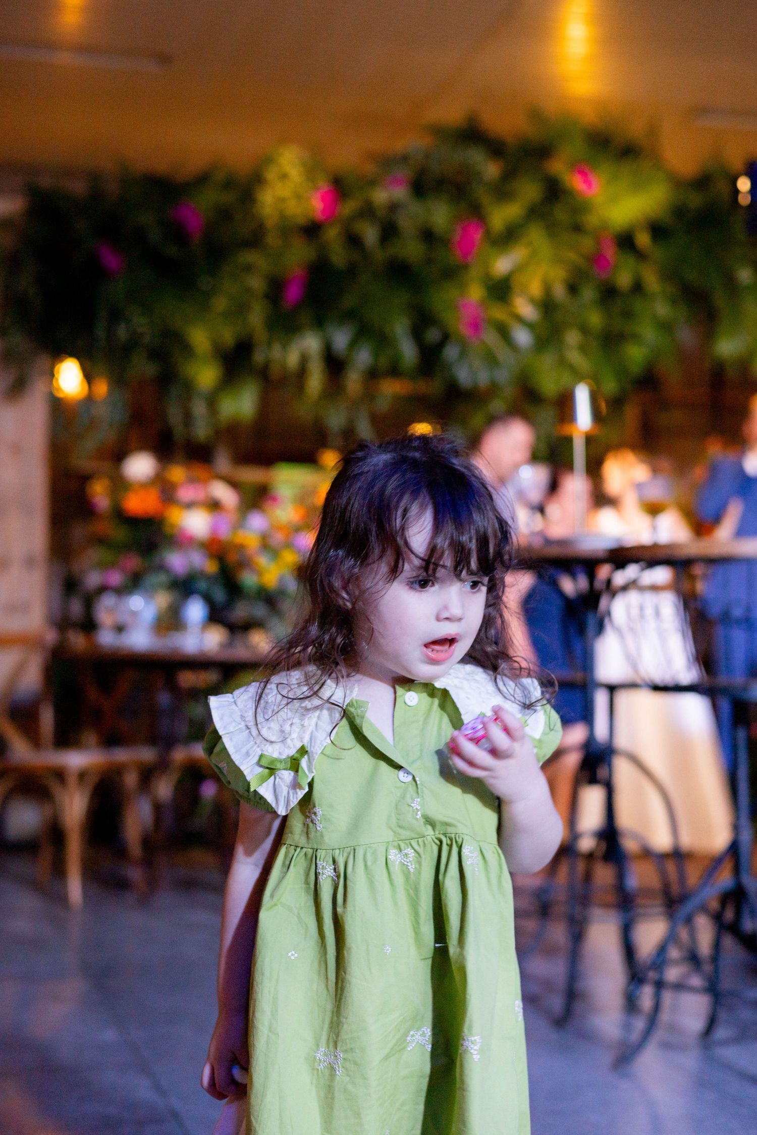 Young girl in green dress holding a flower in a decorated restaurant with lush plants and pink blooms.