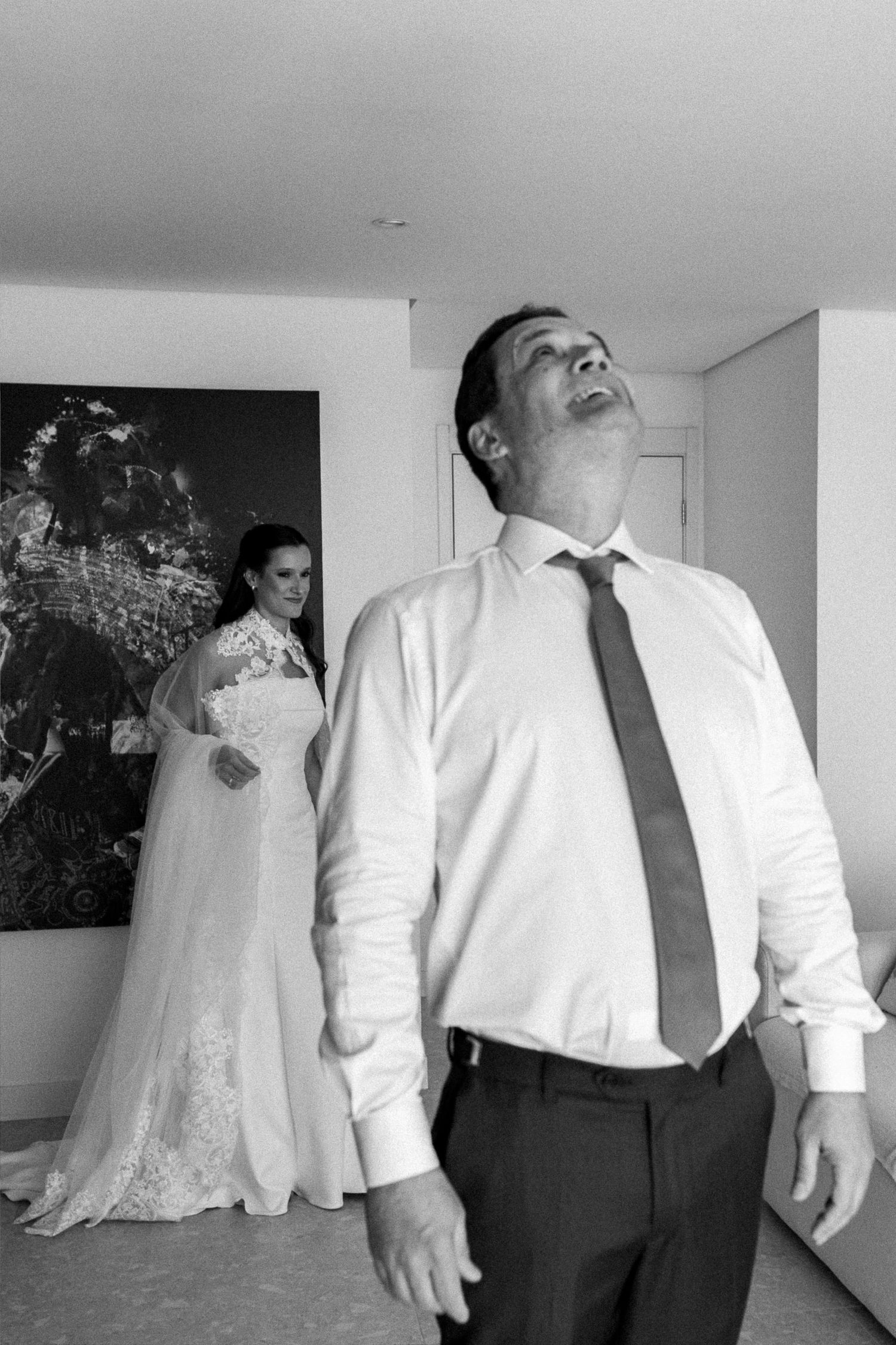 Black and white photo of groom reacting emotionally during first look as bride in lace wedding gown stands behind him.