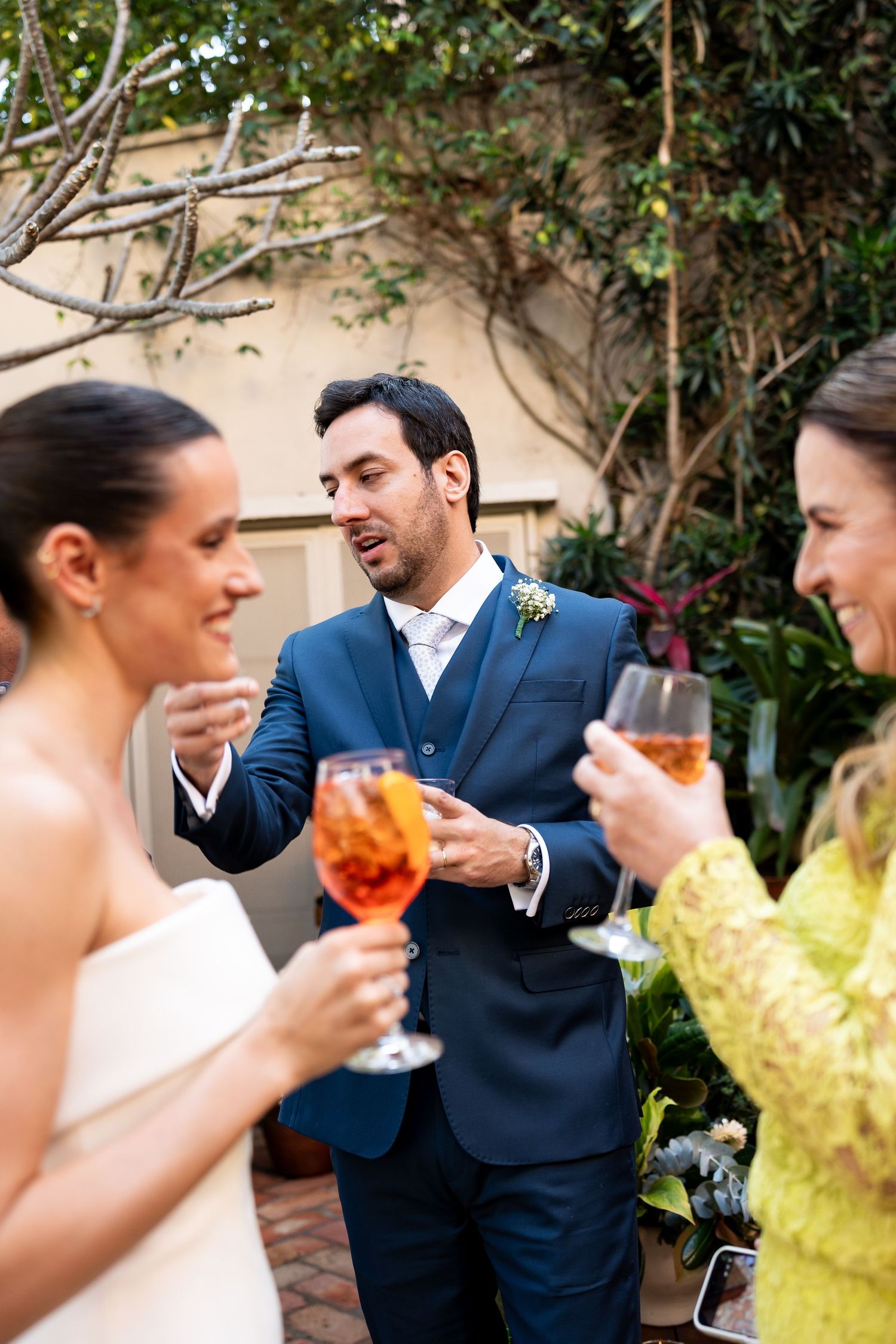 Bride and groom toasting with Aperol Spritz cocktails alongside a guest in yellow at an outdoor wedding reception.