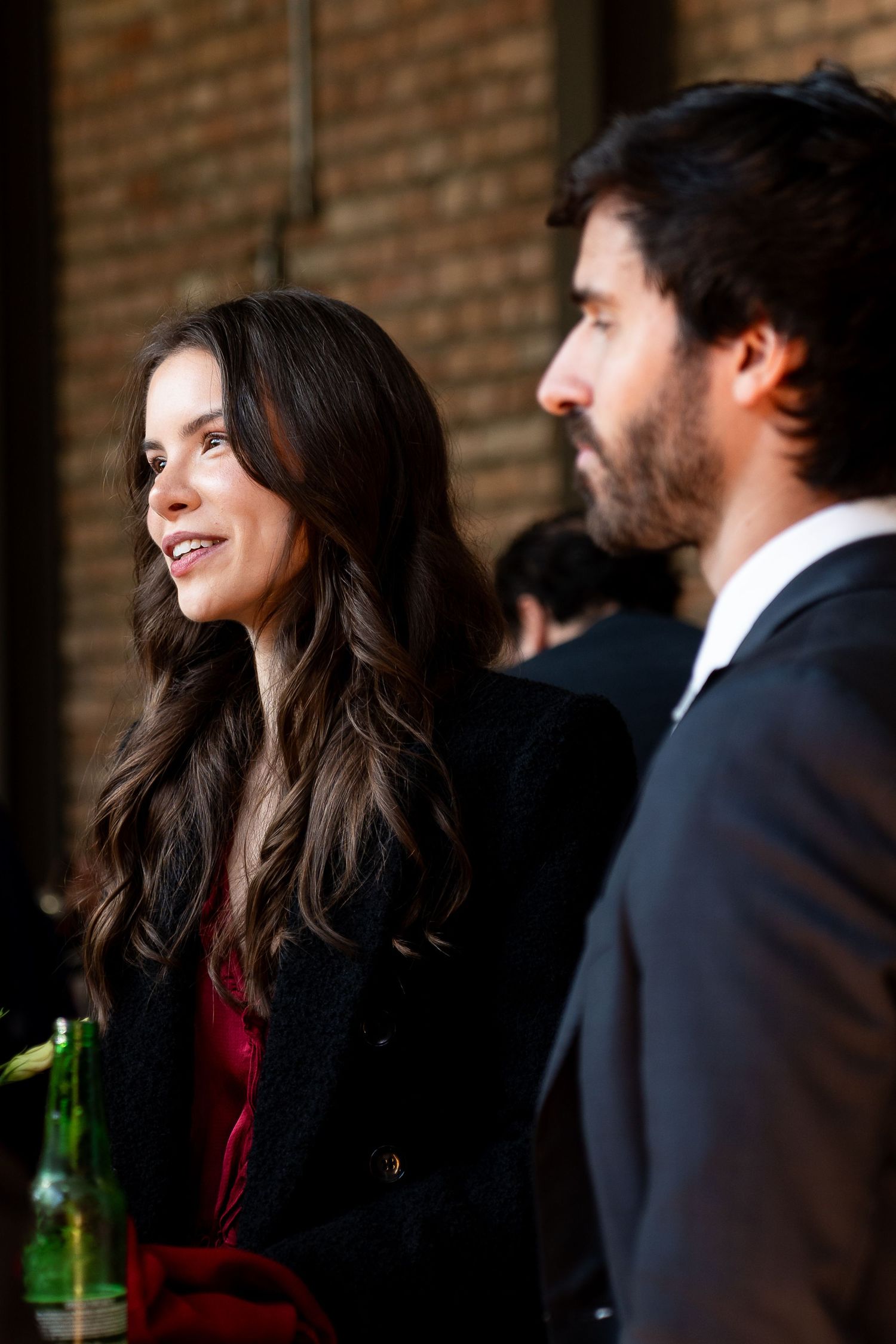 A woman with long dark hair in a black coat smiles at a man in a suit at an indoor social event.