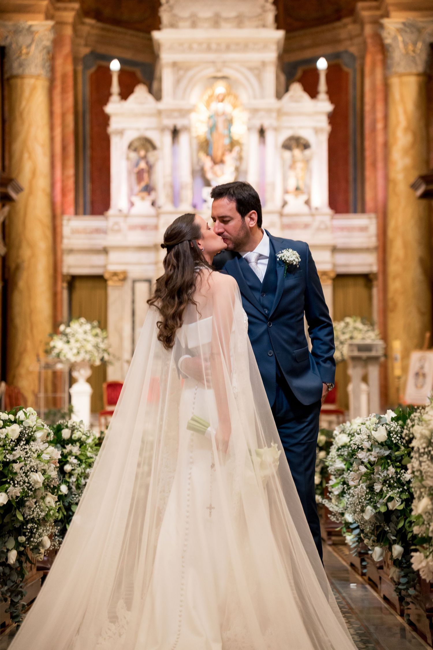 Bride and groom share a first kiss at the altar of an ornate church, surrounded by white floral arrangements.