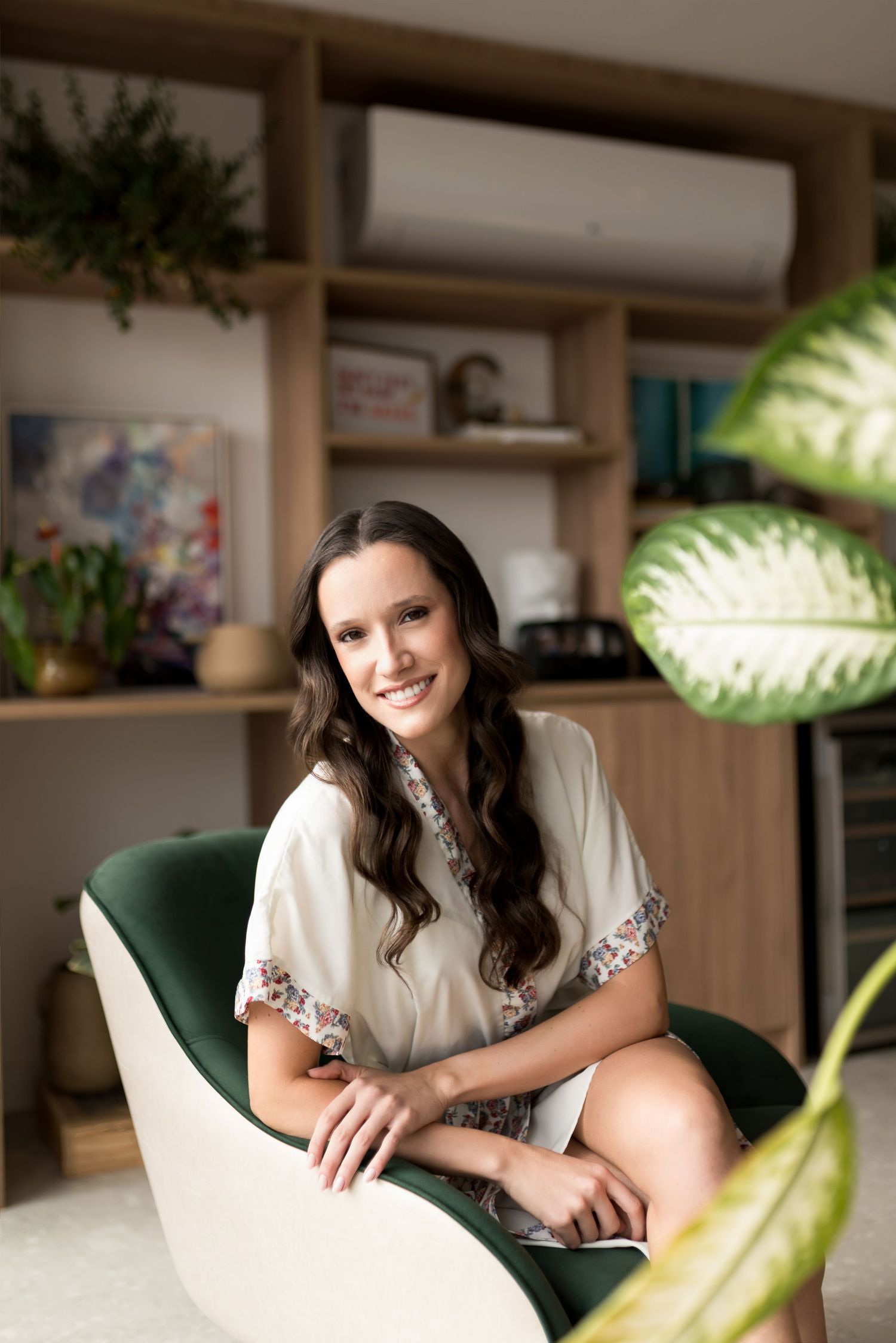 Smiling young woman with long dark hair sitting in a green velvet chair in a stylish home office with wooden shelves.