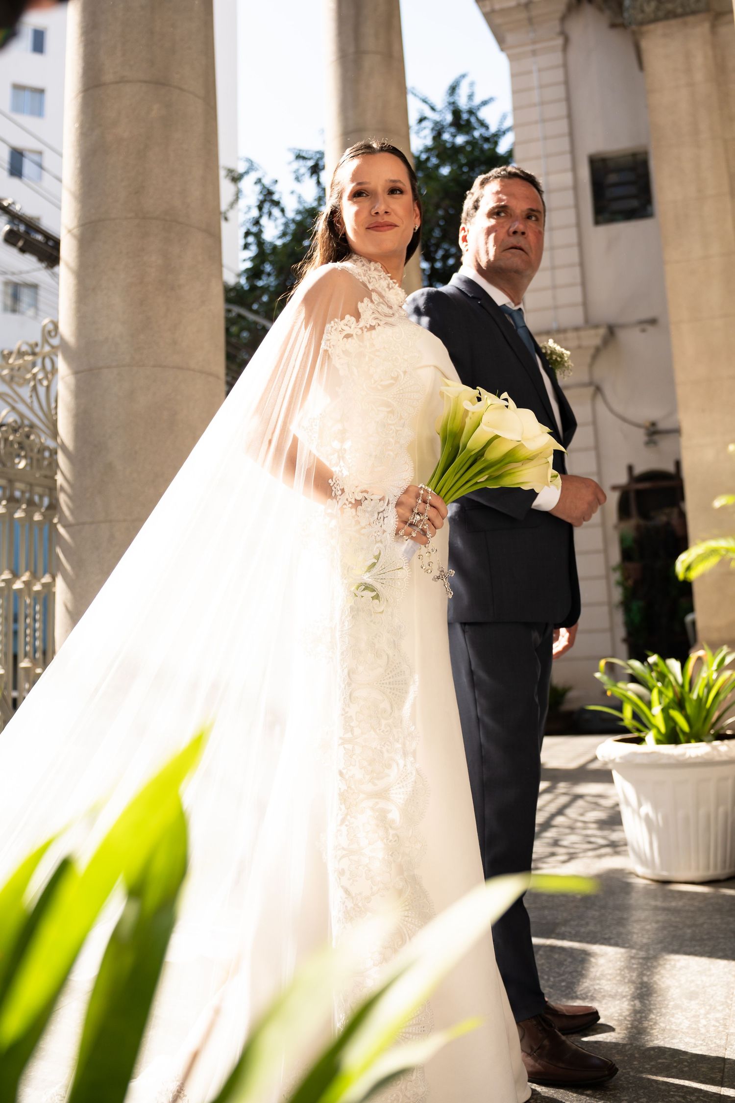 Bride in white lace gown with veil holds calla lily bouquet standing with groom in dark suit outside church.