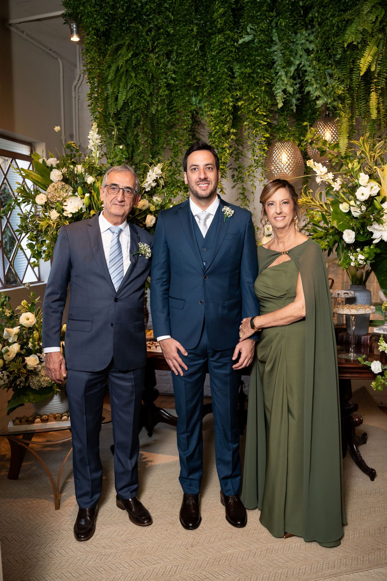Three elegantly dressed people pose together at a wedding reception surrounded by lush green floral arrangements.