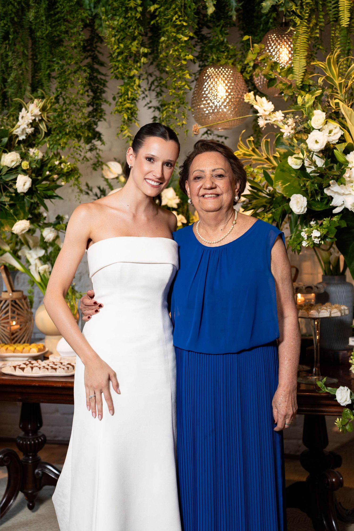 Bride in white strapless gown poses with older woman in blue dress at elegant floral wedding reception backdrop.