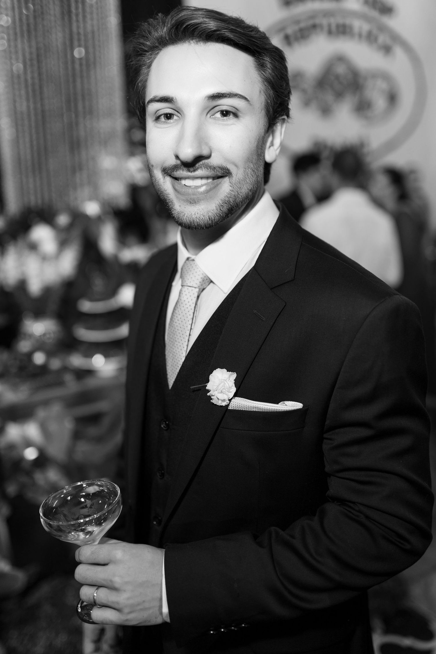 Elegant young man in black suit with boutonniere holds champagne glass at a formal wedding reception.