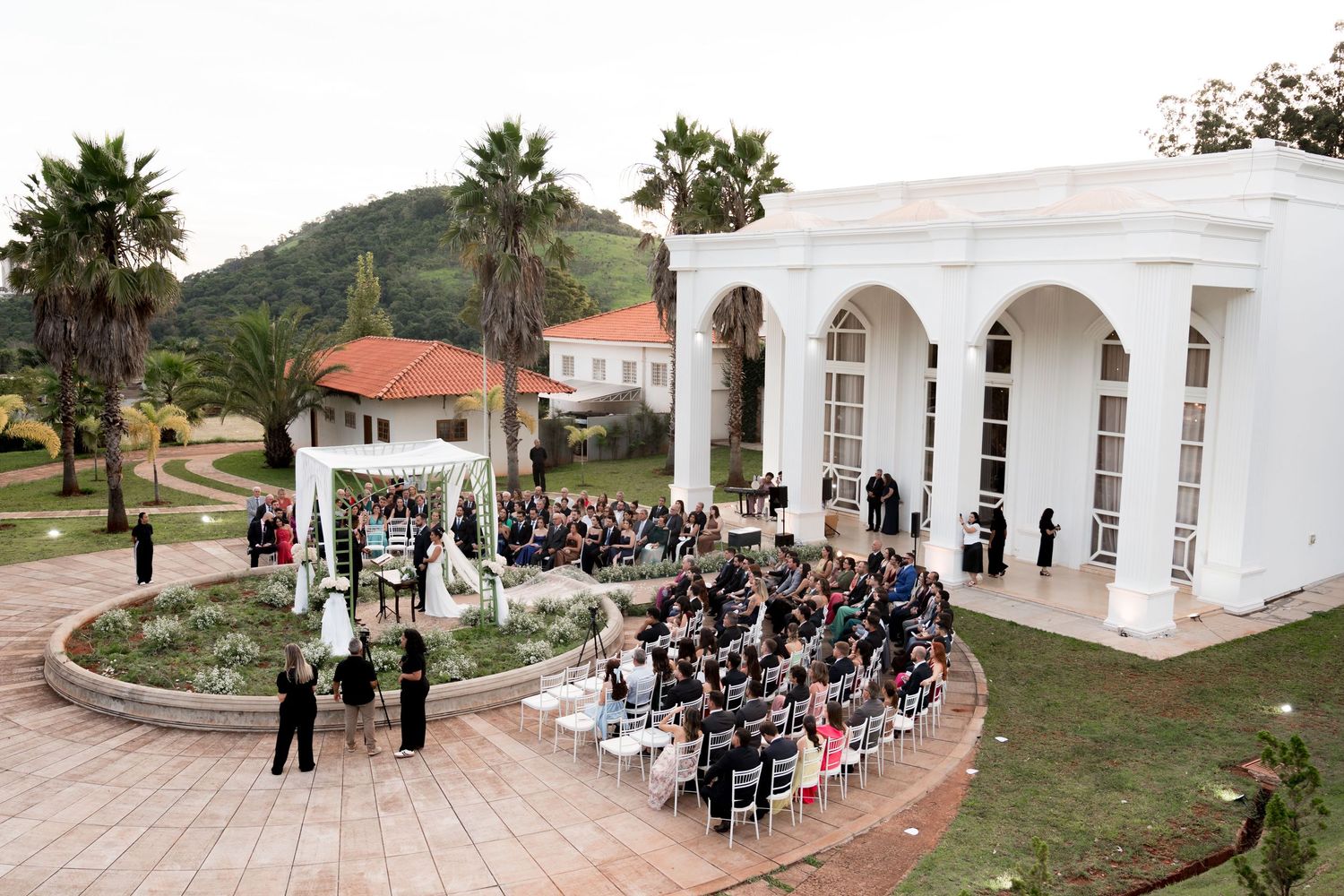 Aerial view of elegant outdoor wedding ceremony at white columned venue surrounded by palm trees.