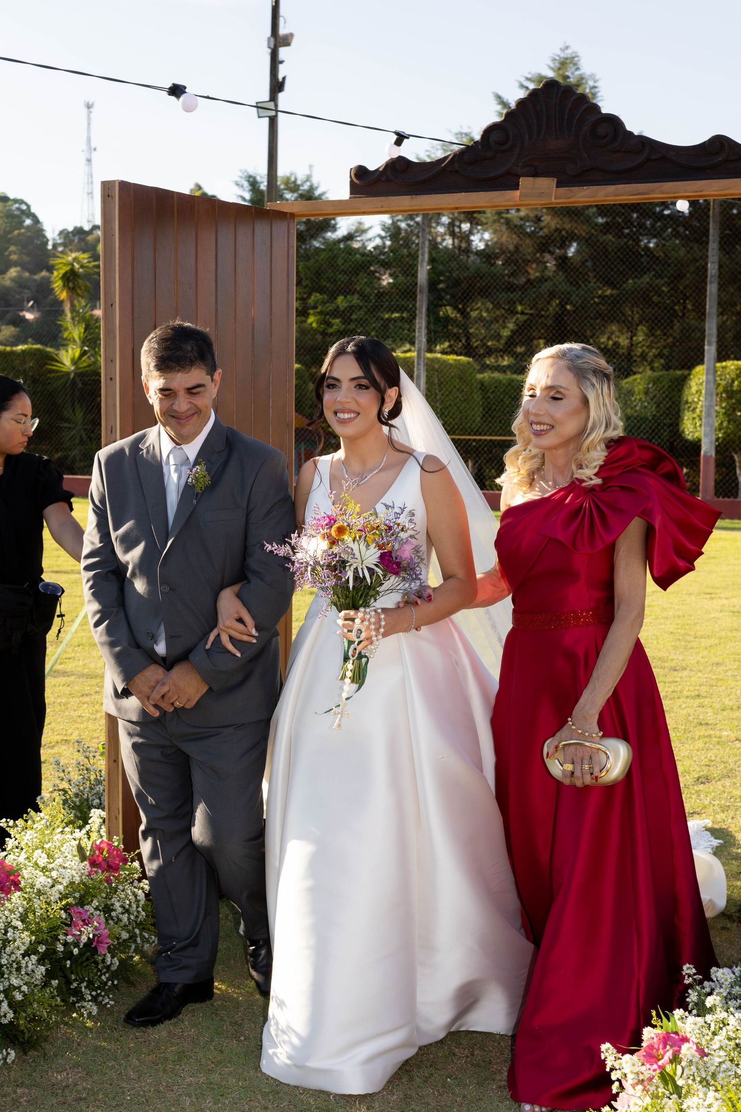 Bride in white gown with bouquet walks with man in gray suit and woman in red dress at outdoor wedding ceremony.
