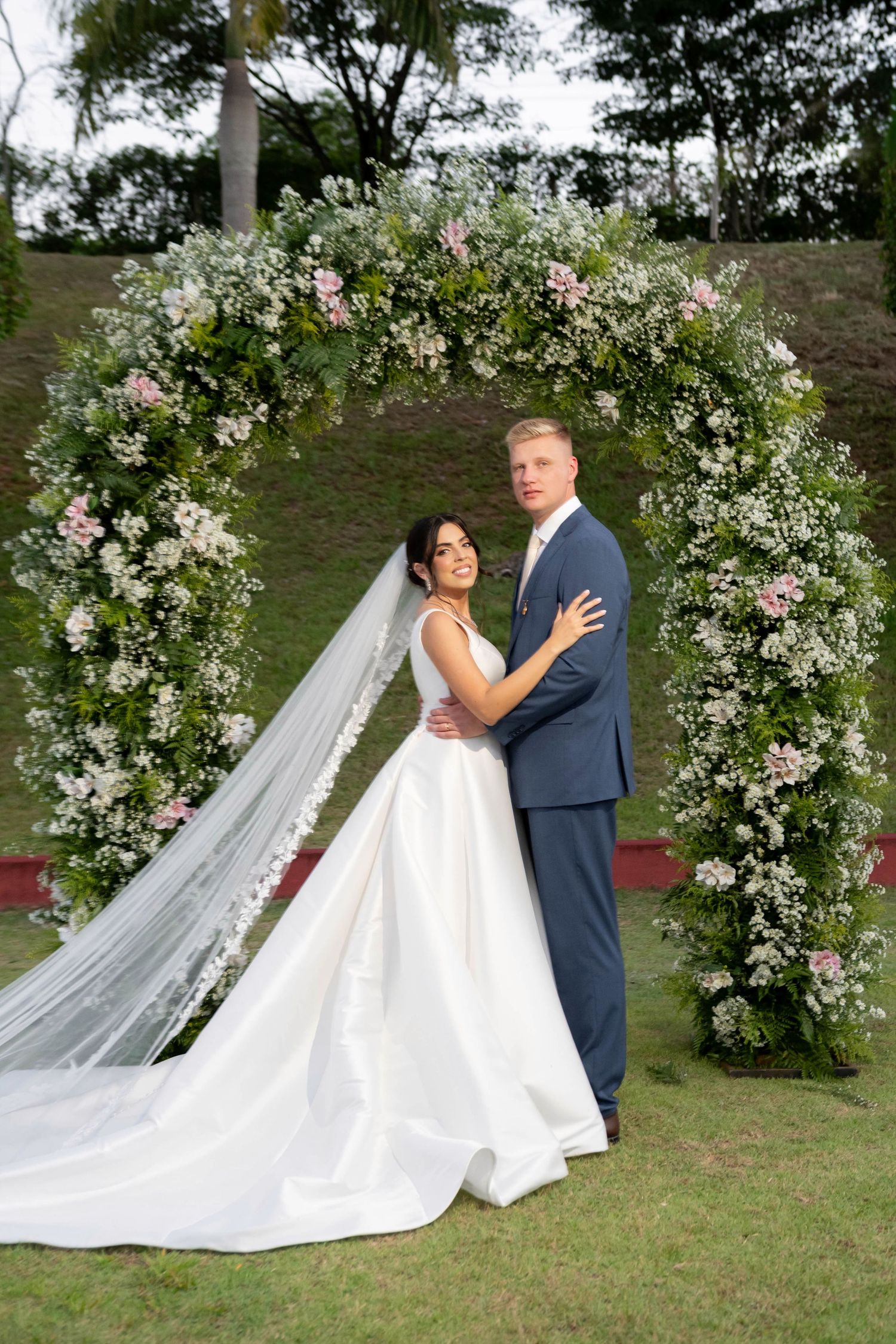Bride and groom pose under a floral arch with white and pink flowers on a green lawn.