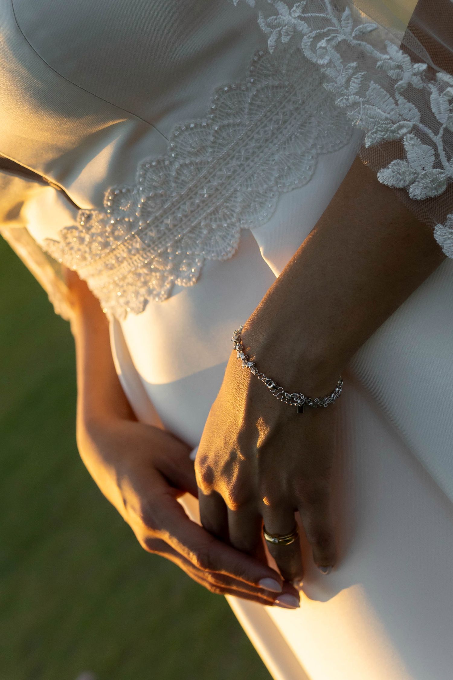 Close-up of bride's hands clasped together, wearing a diamond bracelet and ring, against a white lace wedding gown.