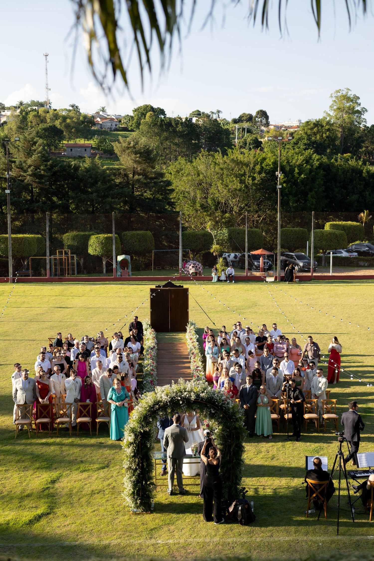 Outdoor wedding ceremony on lush green lawn with floral arch, seated guests, and tree-lined hills in background.