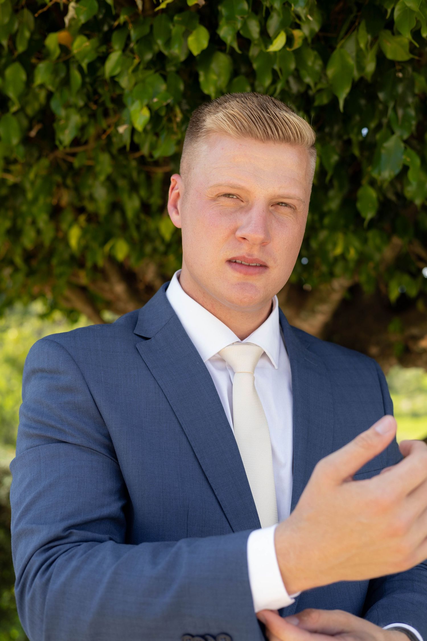 Young blond man in blue suit and white tie adjusting cufflinks outdoors under green tree foliage.